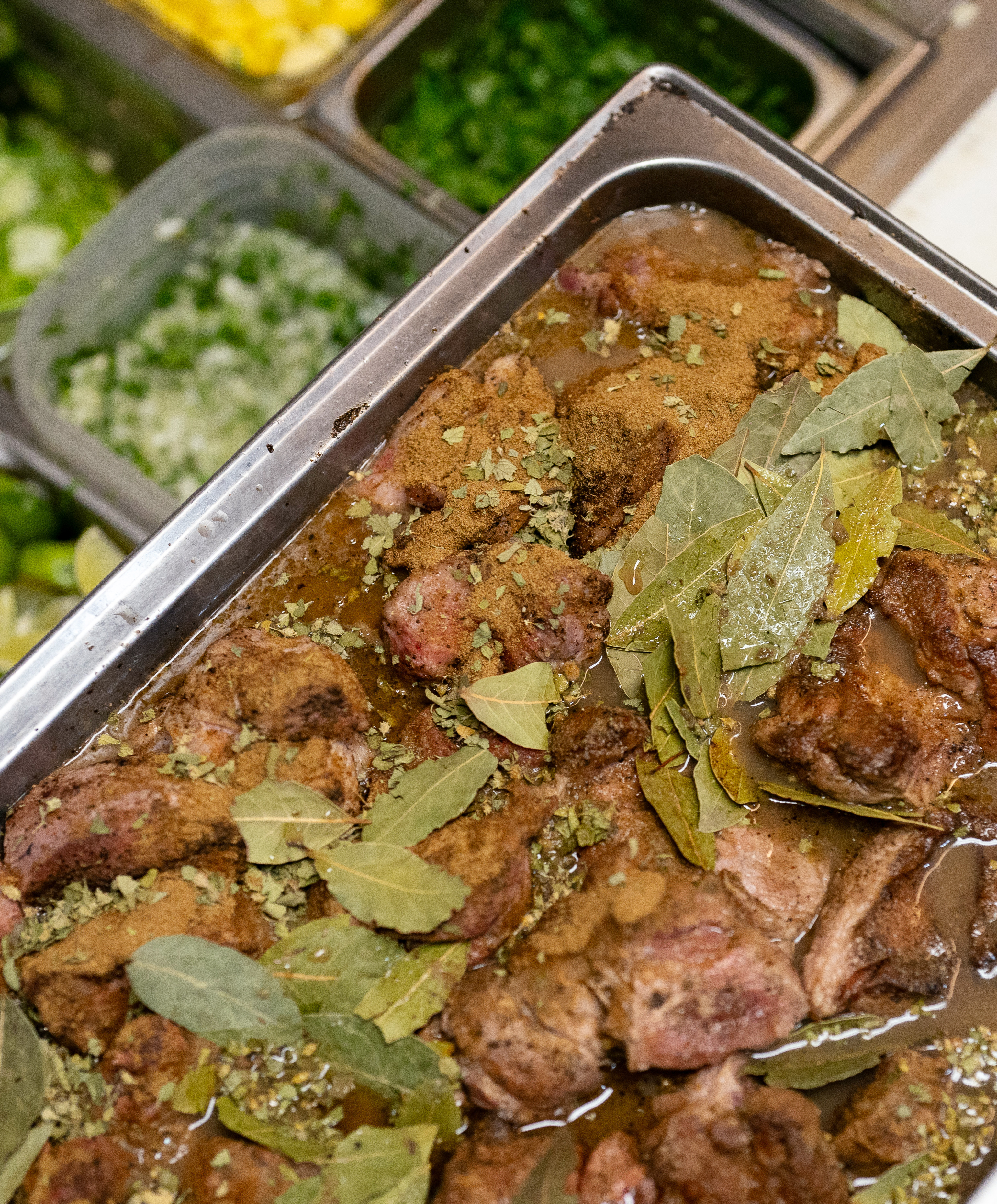 The image displays a tray of seasoned meat pieces topped with bay leaves. In the background, there are containers with chopped vegetables, including green herbs and yellow bell peppers.