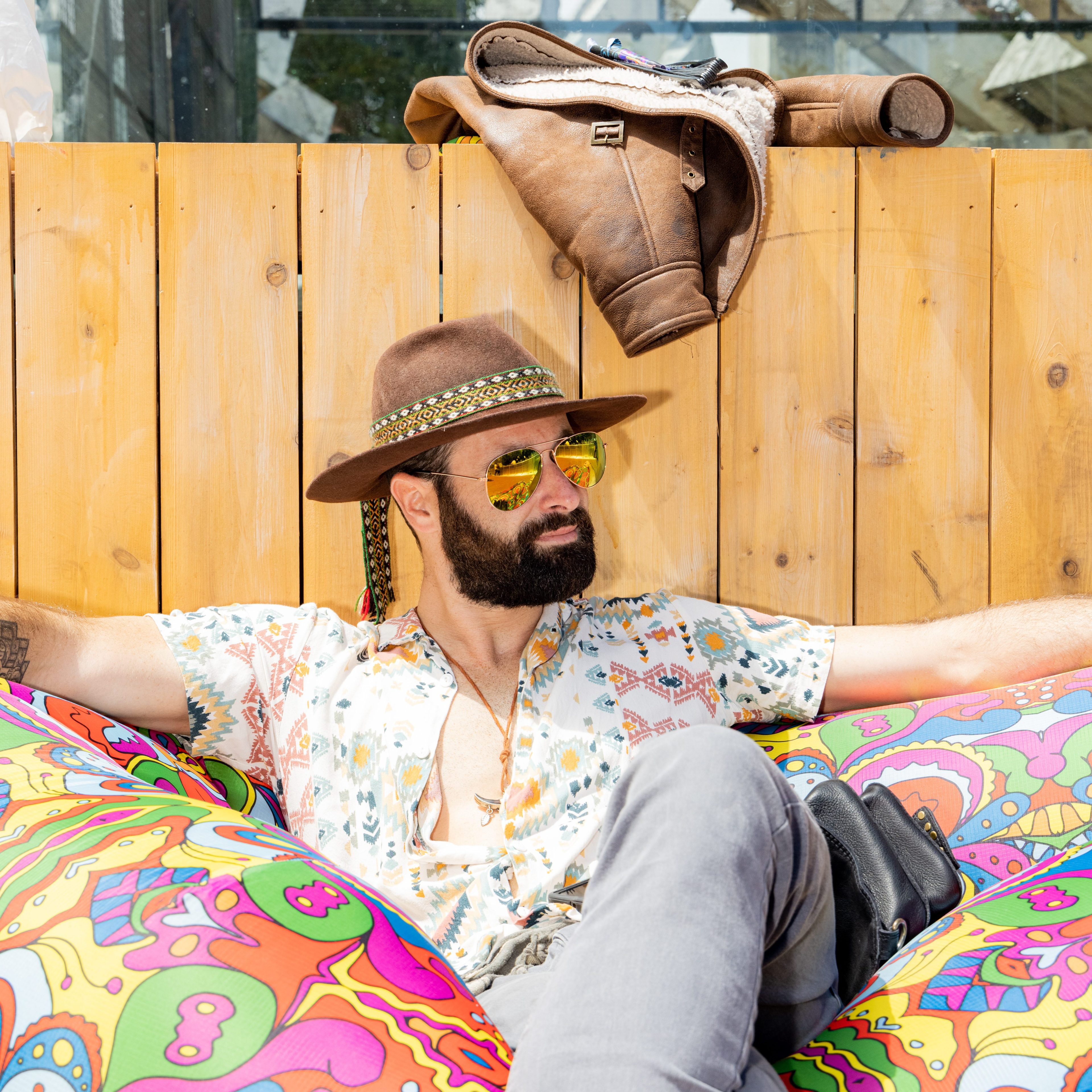 A bearded man wearing a hat, aviator sunglasses, and a patterned shirt lounges on a vibrant, colorful cushion with a wooden fence and jacket hung behind him.