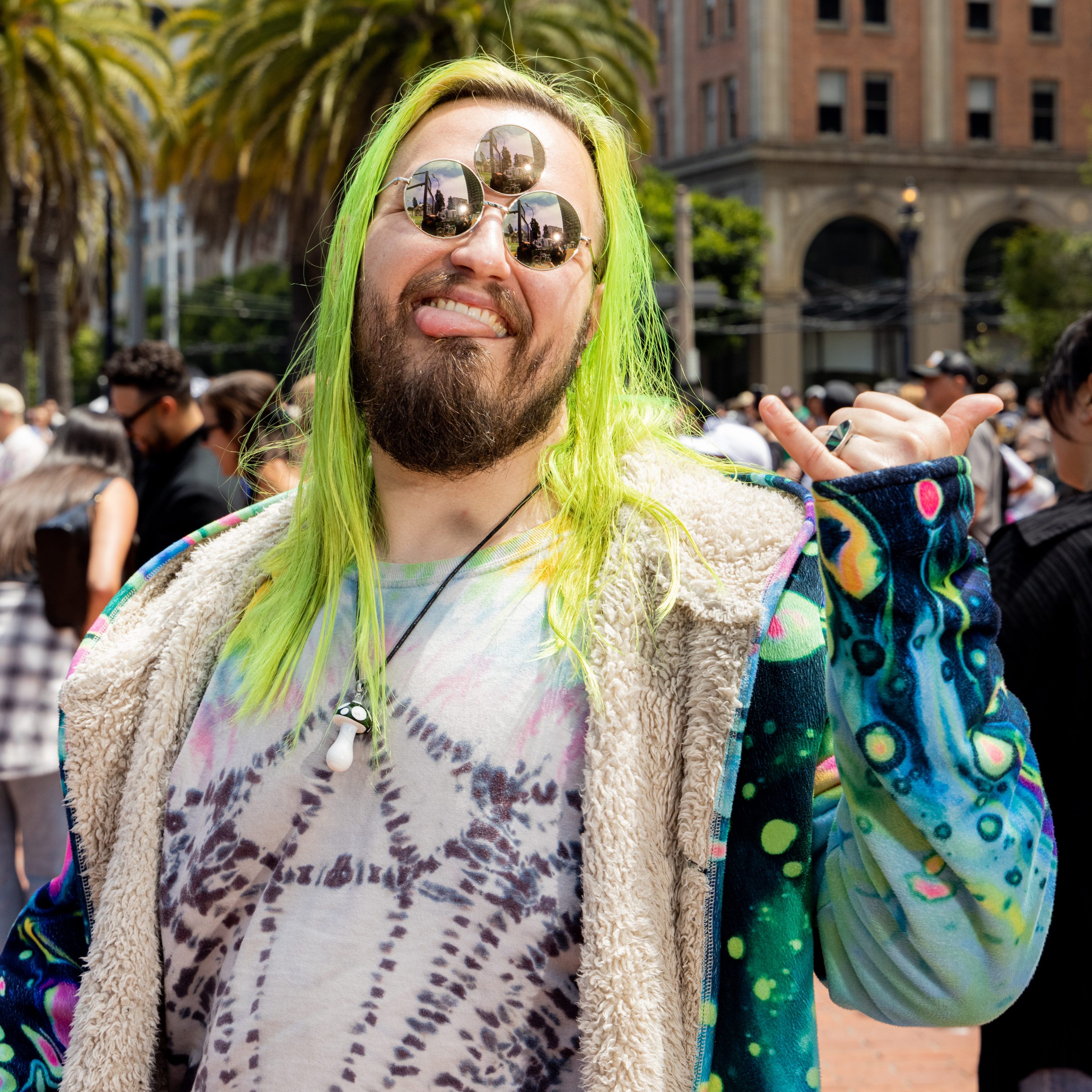 A person with bright green hair, multiple round sunglasses, and a colorful outfit is smiling and sticking out their tongue at an outdoor event with palm trees and buildings in the background.