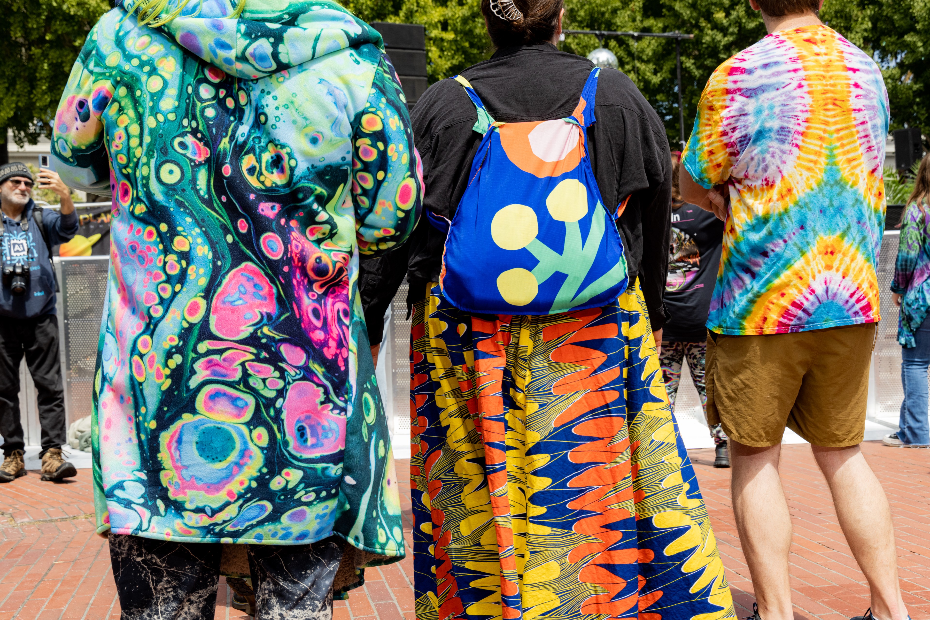 Three individuals wearing colorful, psychedelic-patterned clothing stand outside. One has a tie-dye shirt, another has a vibrant dress, and the last wears a vivid coat.