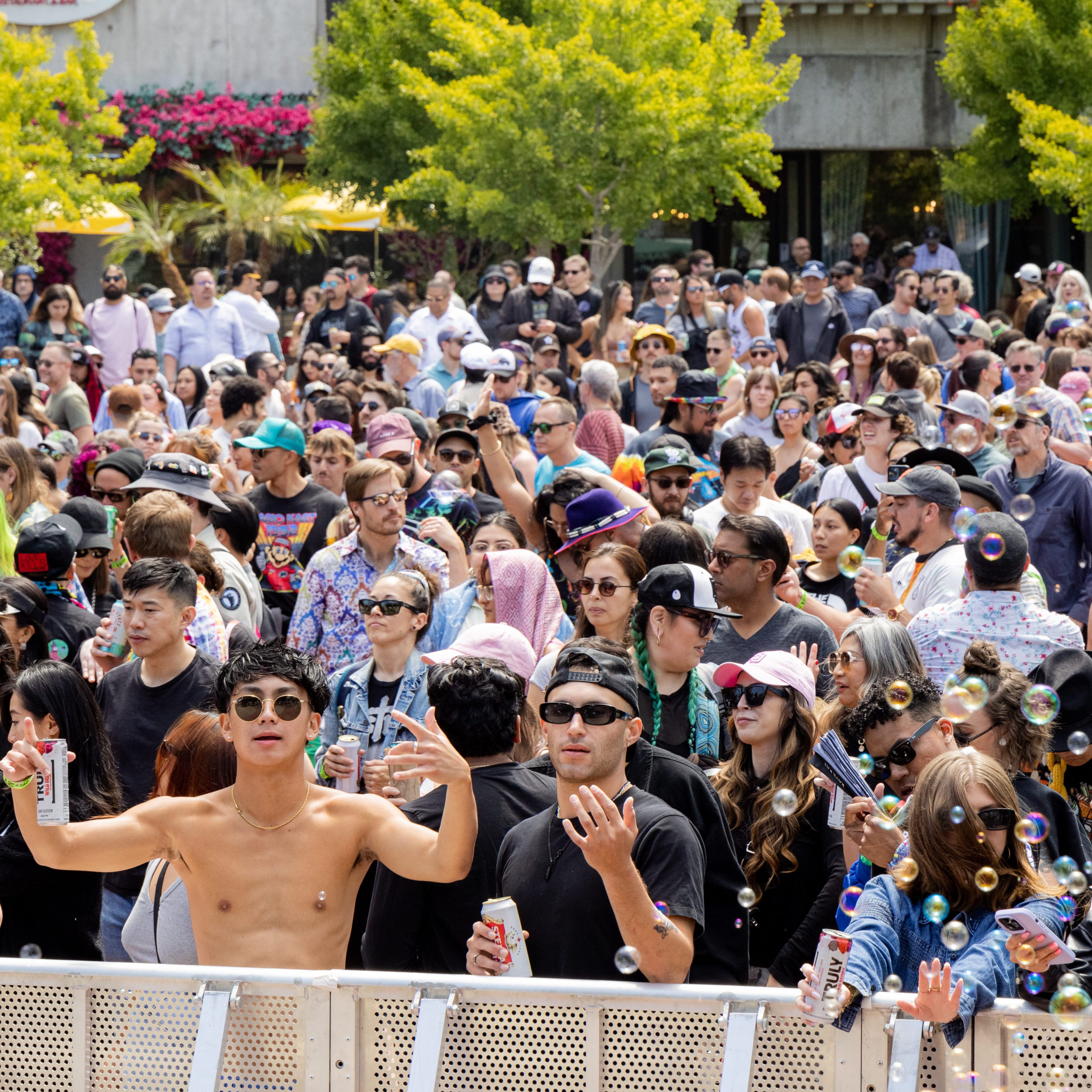 A large, lively crowd gathers at an outdoor event. People wear casual attire, sunglasses, and hats, holding drinks and phones, with bubbles floating around. Trees are in the background.