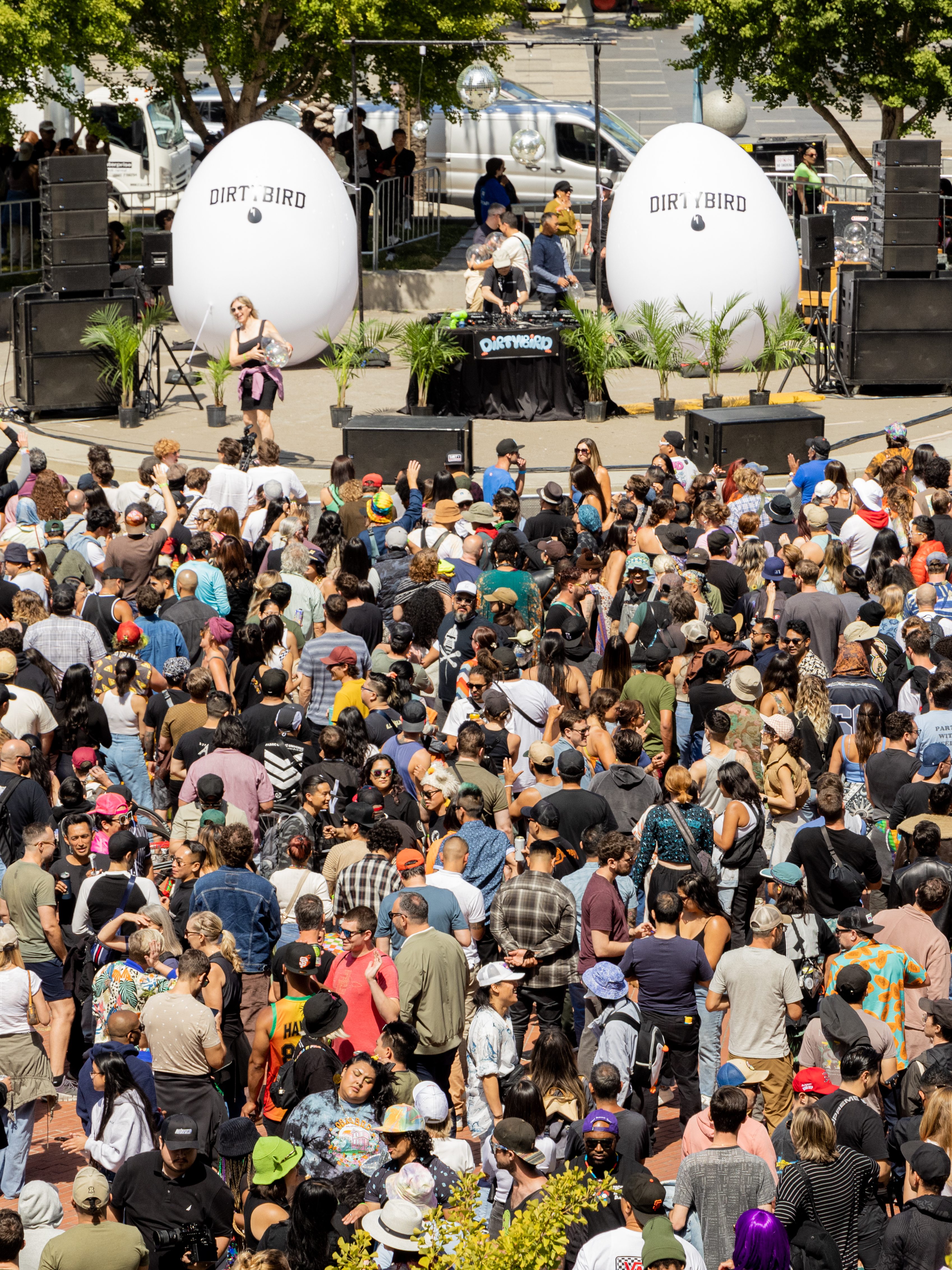 A large crowd gathers outdoors for an event with music. Two giant egg props labeled &quot;DIRTYBIRD&quot; and a DJ booth are on stage, surrounded by greenery and speakers.