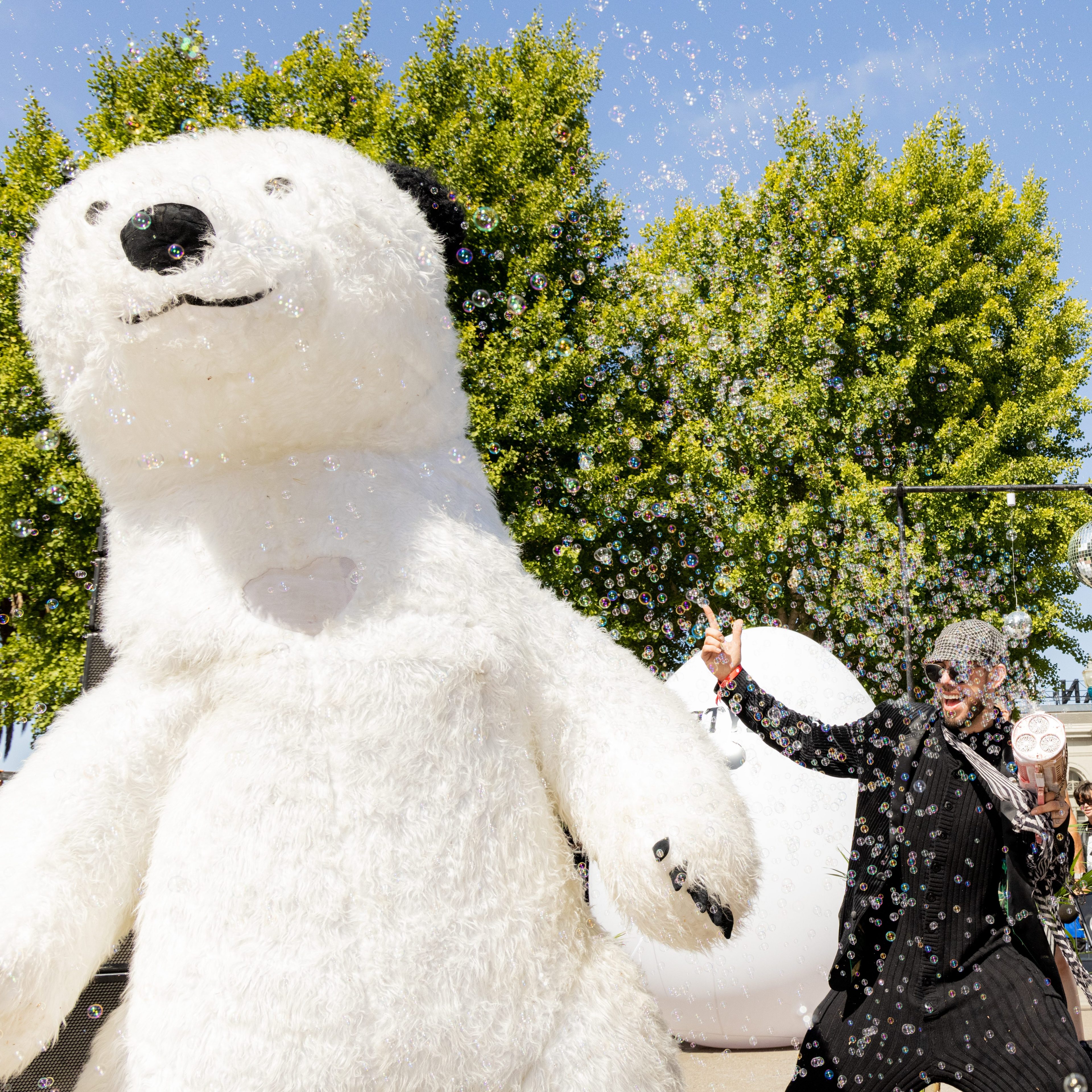 A man in dark clothing happily blows bubbles next to a large, white polar bear costume, with a DJ performing under a blue sky and green trees in the background.