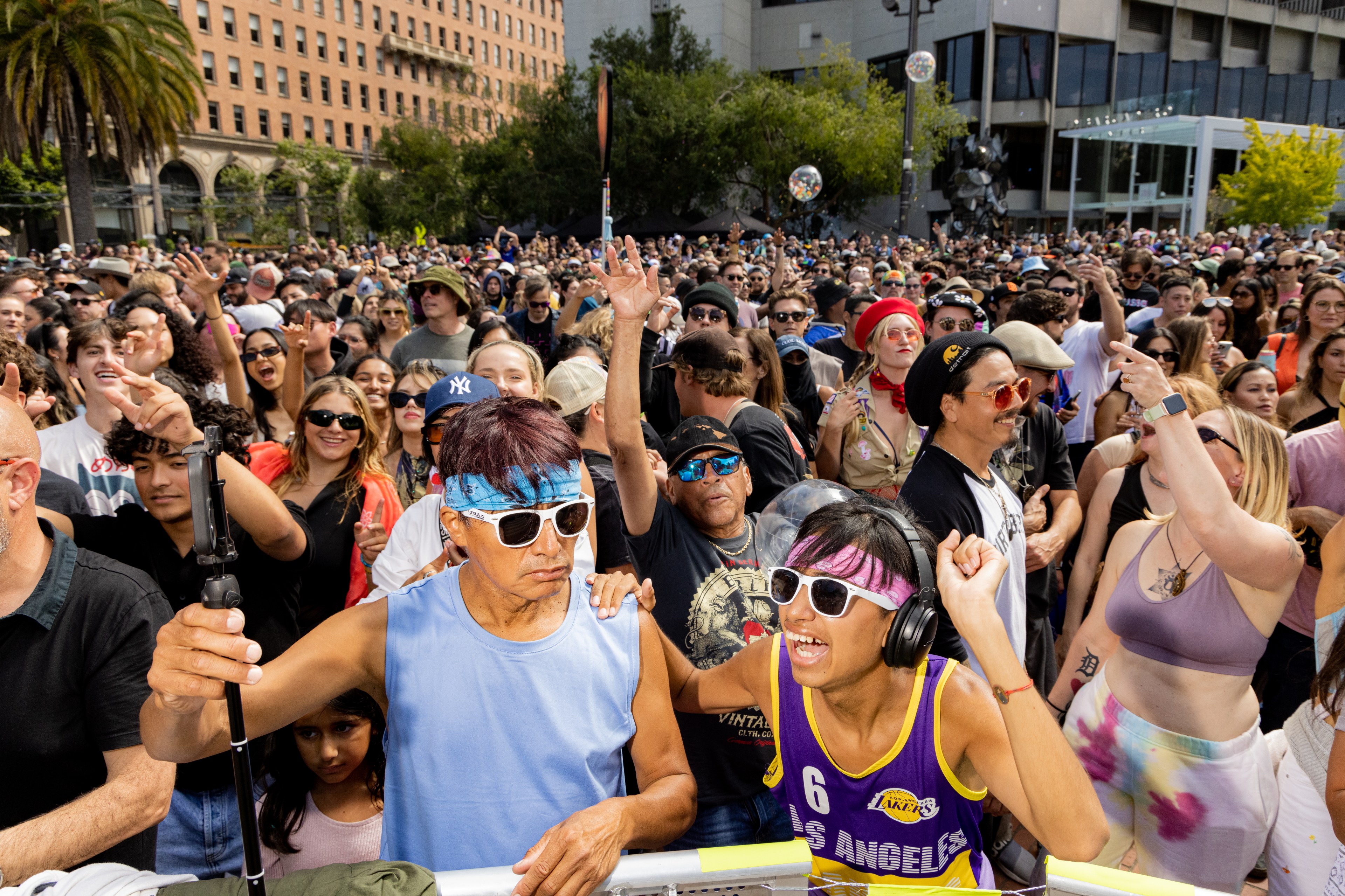 A lively crowd at an outdoor event with people wearing bright clothes, sunglasses, and headbands; some are waving and cheering, with a mixture of joy and energy.