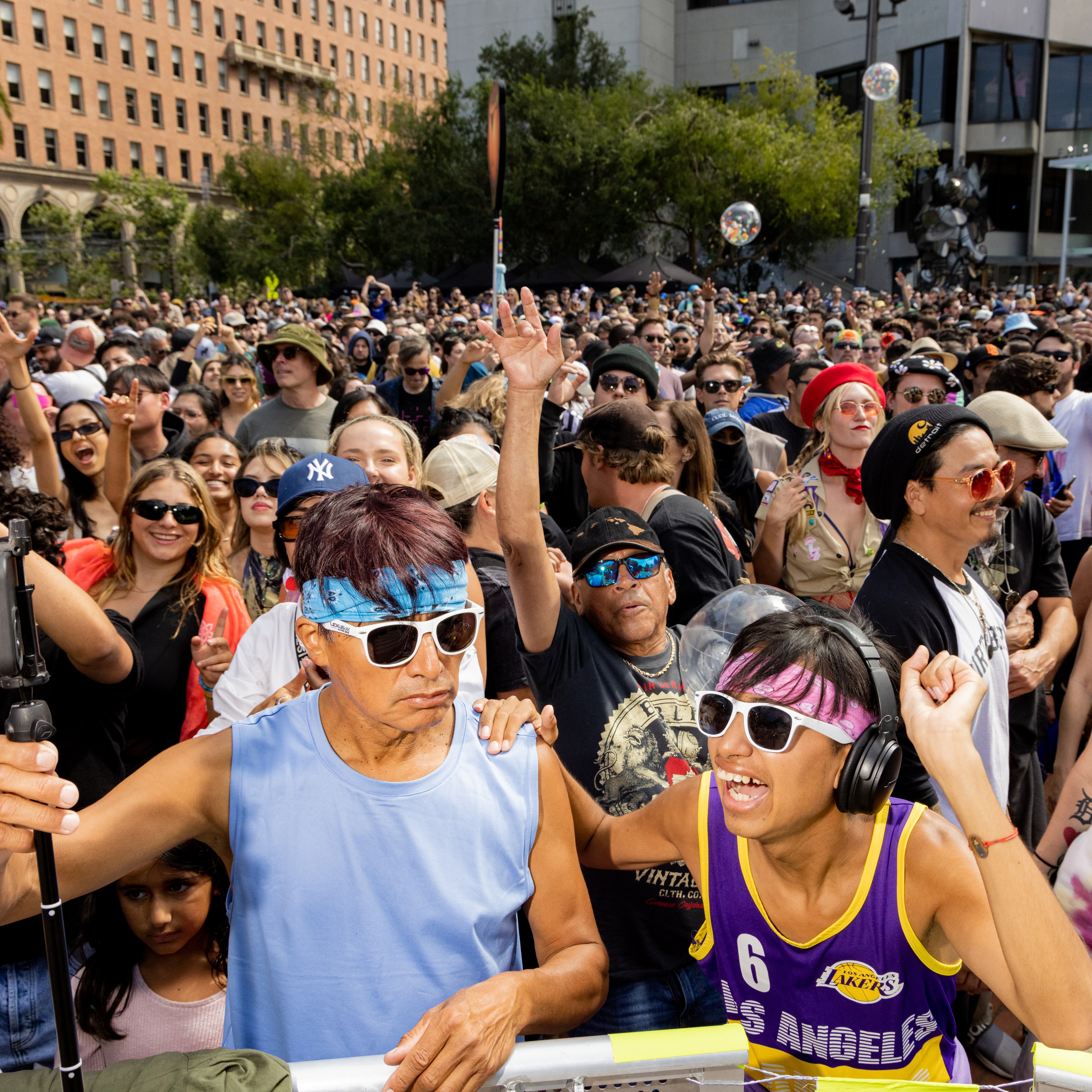 A lively crowd at an outdoor event with people wearing bright clothes, sunglasses, and headbands; some are waving and cheering, with a mixture of joy and energy.