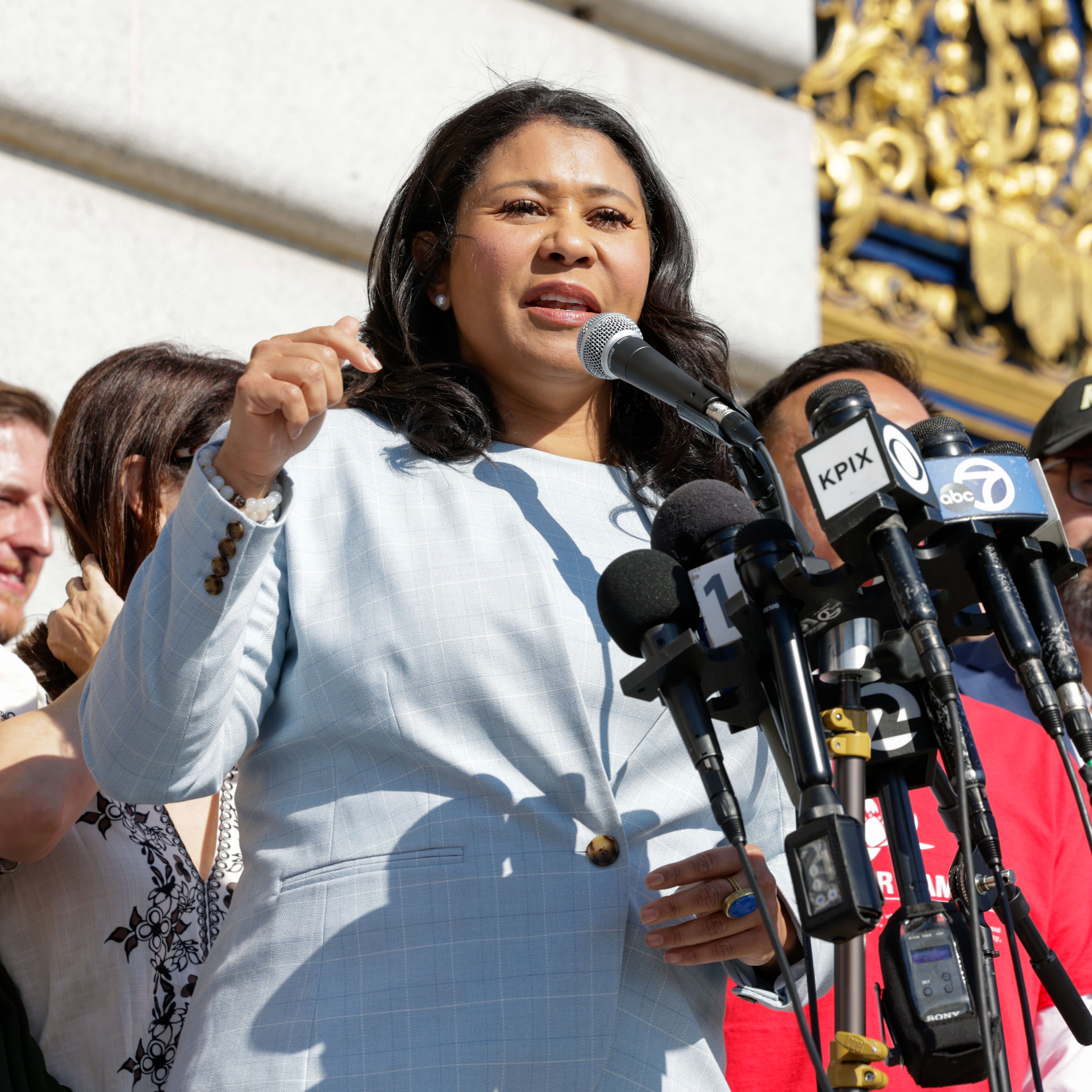 A woman in a light blue suit is speaking at a podium with multiple microphones. She is surrounded by several people, and intricate gold decor can be seen in the background.