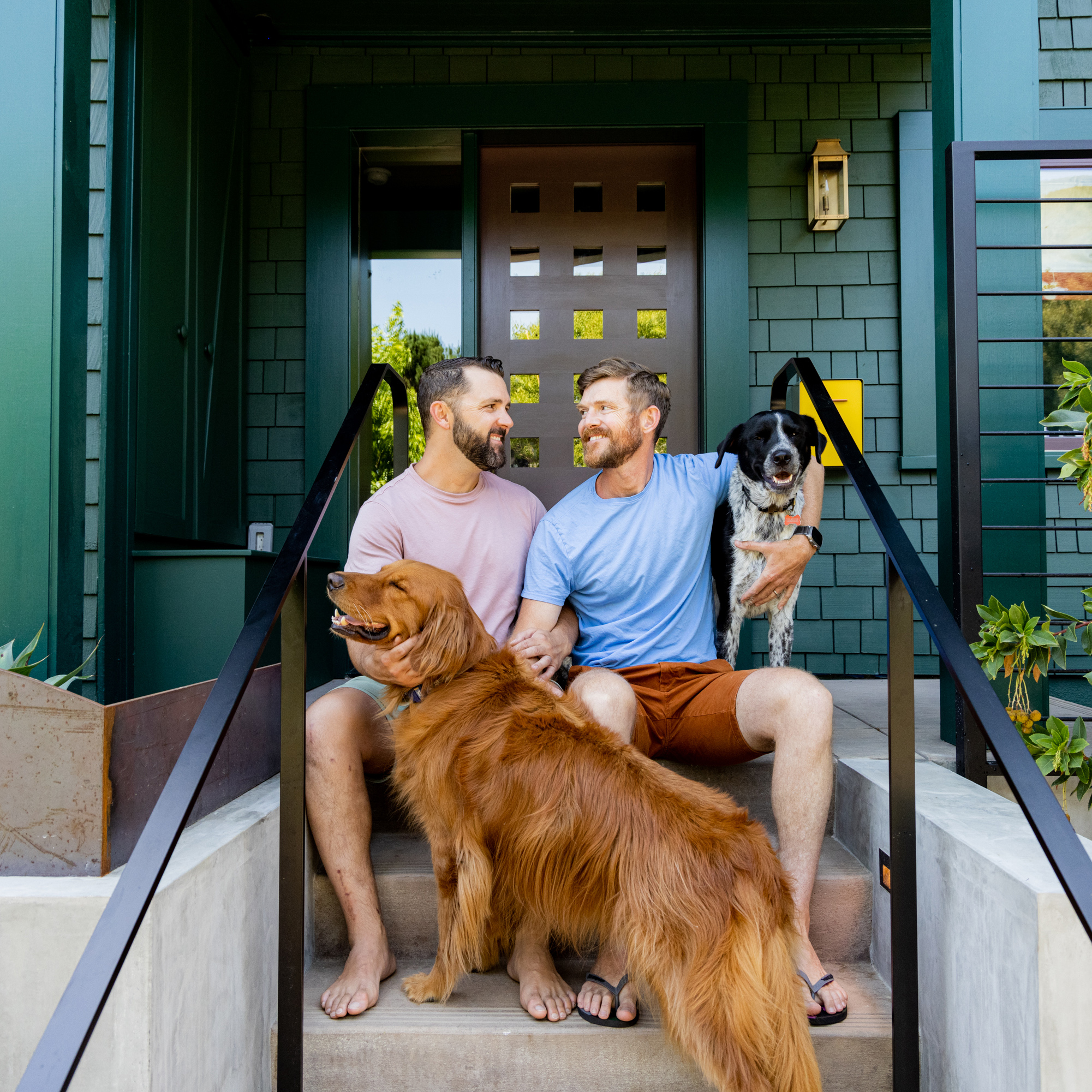 Two men sit on their porch steps, smiling at each other, each petting a dog. One dog is a golden retriever, and the other is a black and white mixed breed.