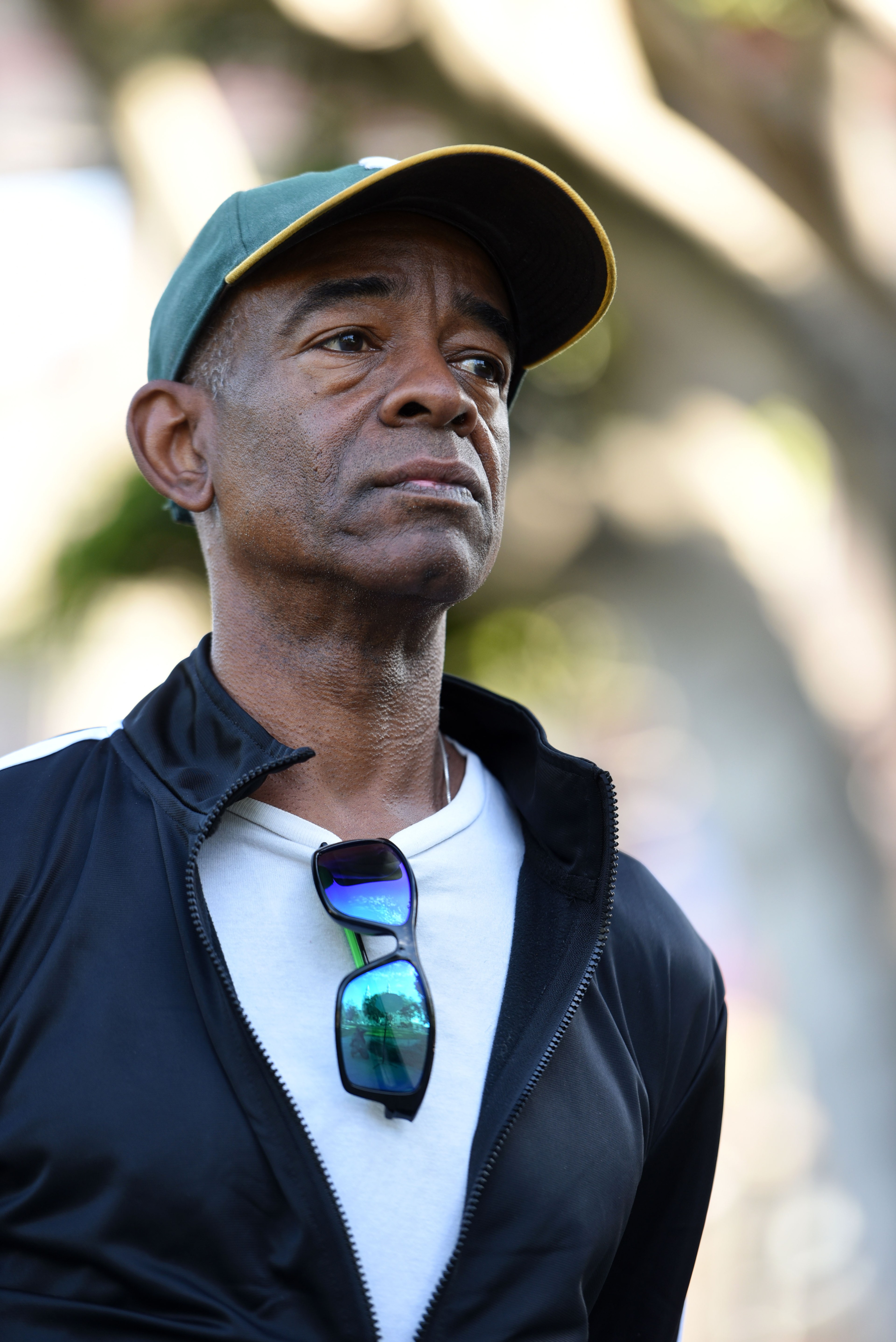 A man wearing a green cap and black jacket looks off into the distance with a serious expression. Blue-rimmed sunglasses hang from the neckline of his white shirt.