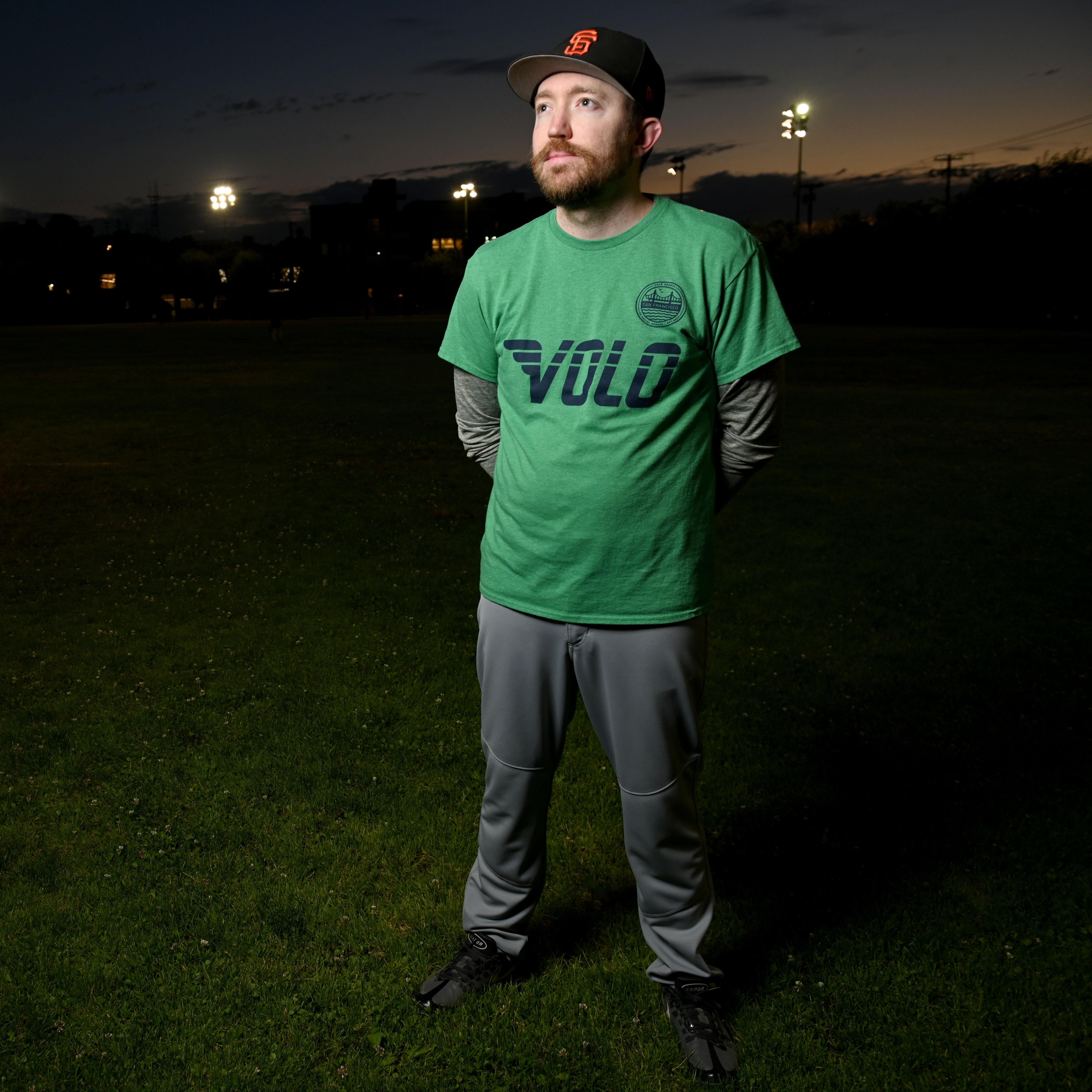 A bearded man in a green &quot;Volo&quot; t-shirt and gray pants stands on a grass field at dusk, wearing a dark baseball cap, with stadium lights in the background.