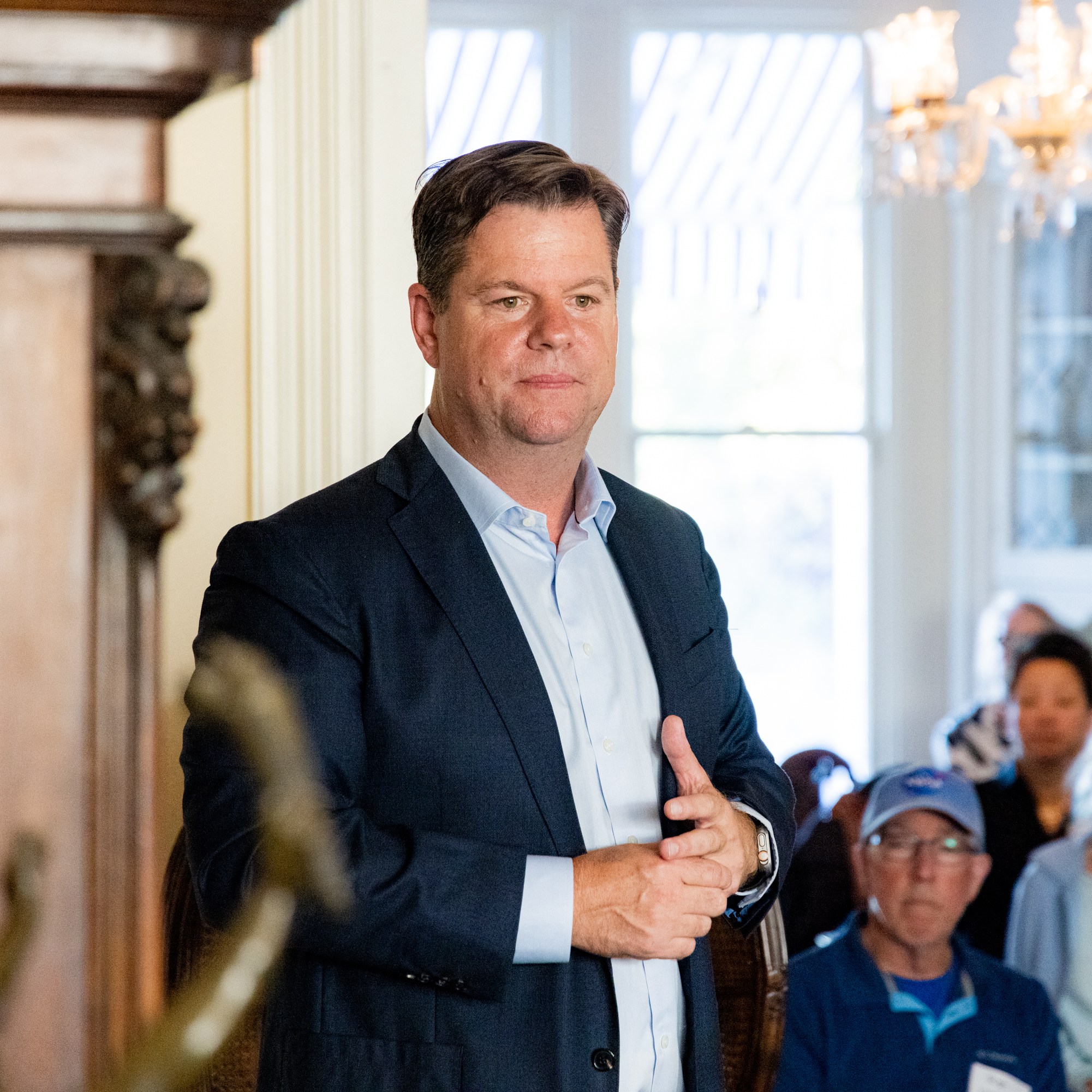 A man in a dark suit and light blue shirt stands in a room with a chandelier. Several seated people, some wearing masks and hats, are attentively watching him.