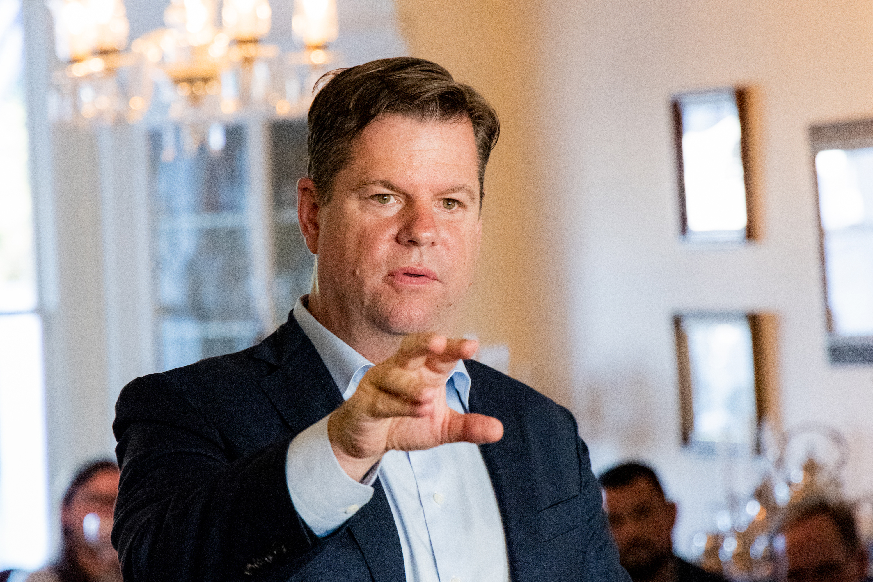 A man in a dark suit jacket gestures with his hand. He appears to be speaking. The background features a chandelier and blurred people.