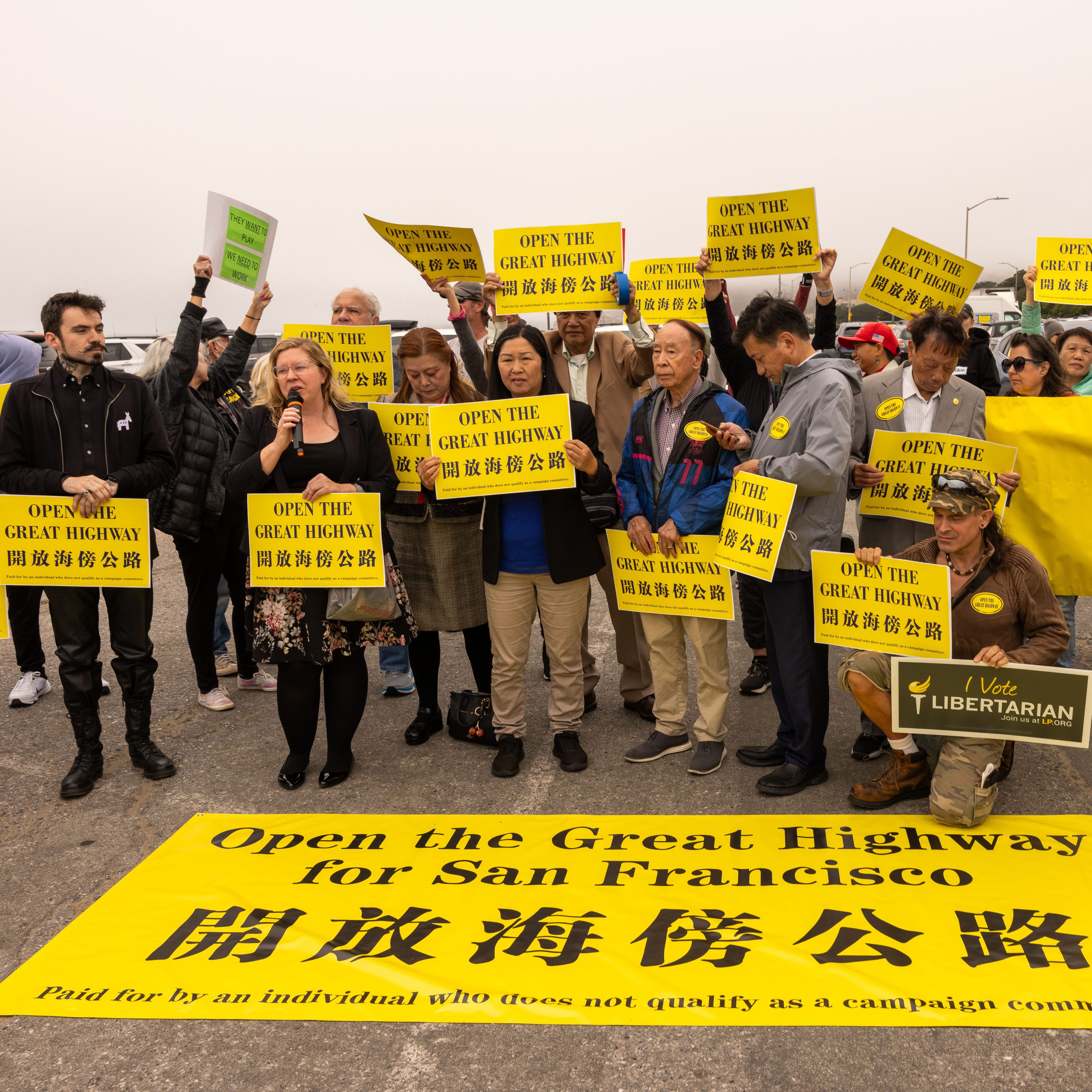 A group of people are gathered holding yellow signs that read &quot;Open the Great Highway&quot; in both English and Chinese. They are participating in a protest or rally.