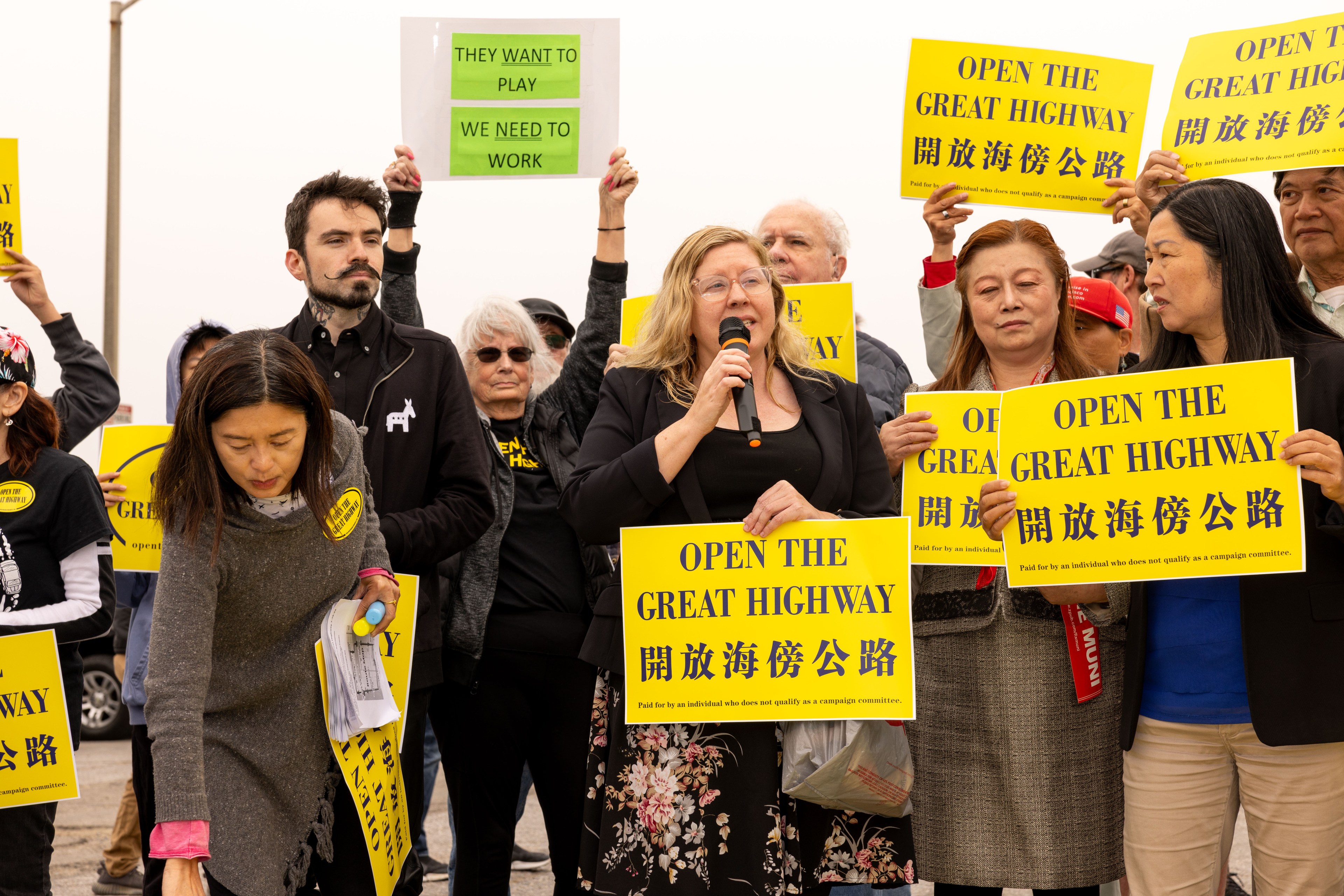 A group of people holds yellow signs saying &quot;Open the Great Highway&quot; in English and Chinese. A woman speaks into a microphone. One sign says, &quot;They want to play, we need to work.&quot;