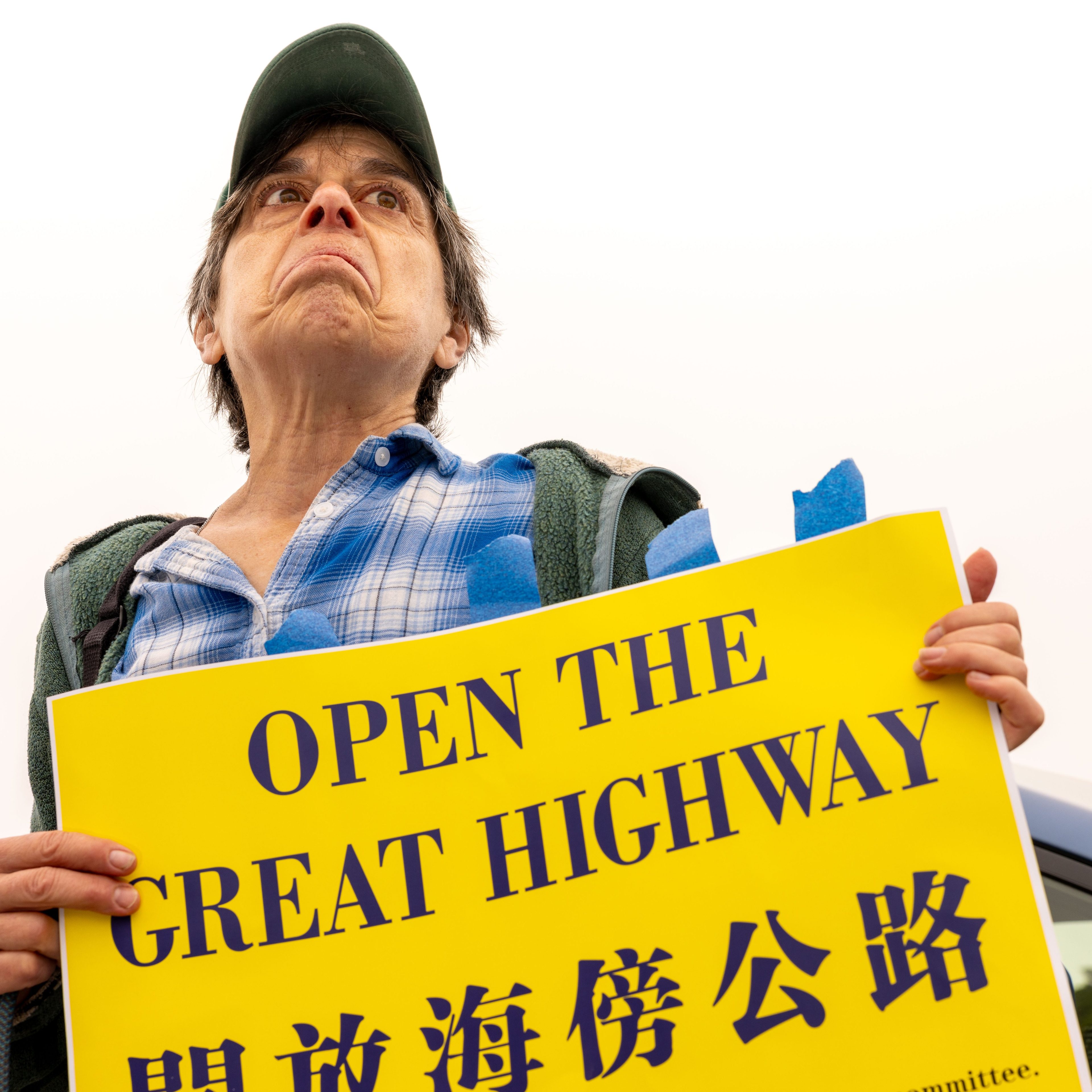 A person in a cap and plaid shirt holds a yellow sign reading &quot;OPEN THE GREAT HIGHWAY&quot; in English and Chinese. Their expression is serious and determined.