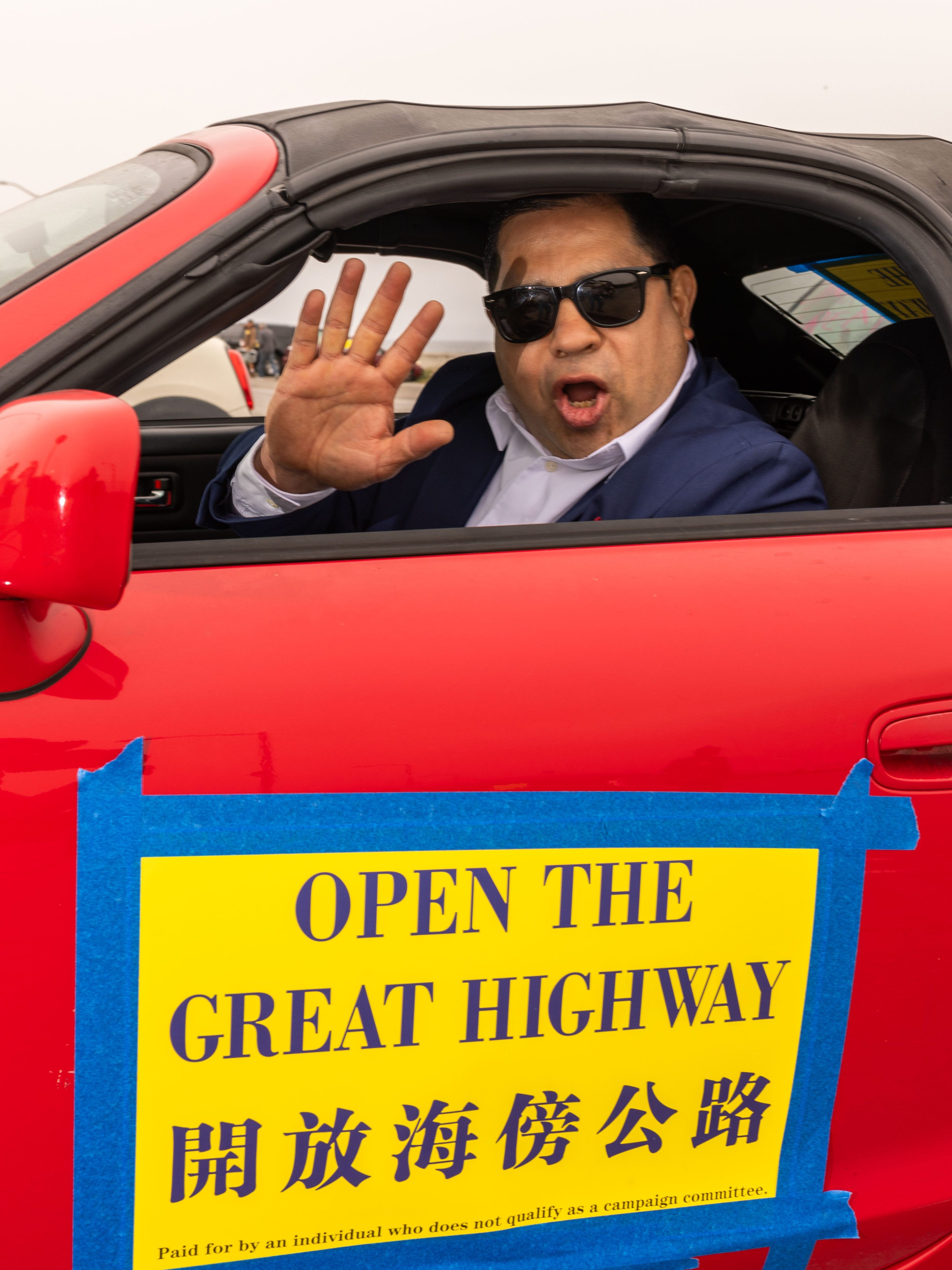 A man in sunglasses and a suit waves and speaks from a red convertible with a sign reading "OPEN THE GREAT HIGHWAY" in English and Chinese taped to the door.