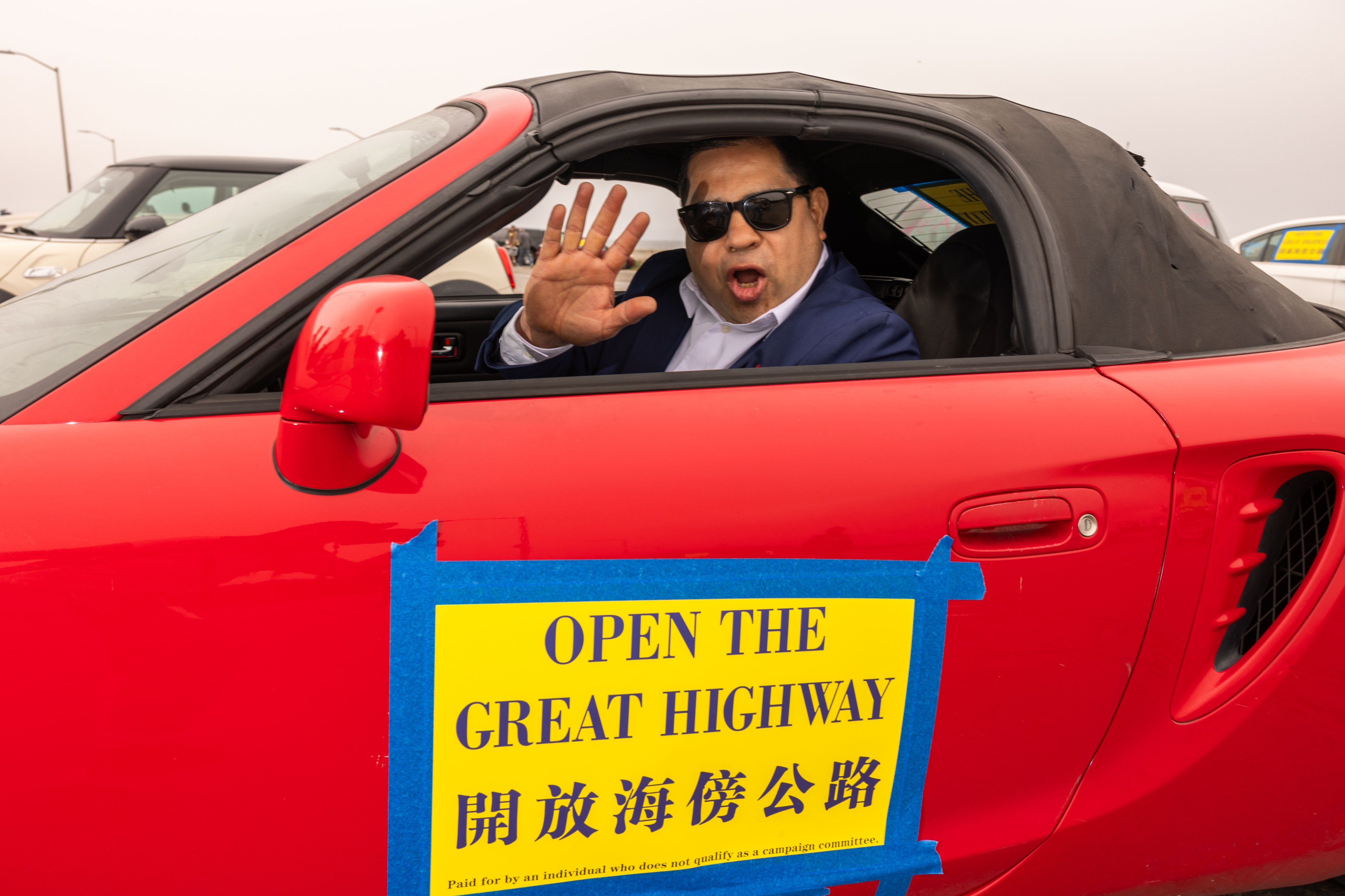 A man in sunglasses inside a red convertible, waving and shouting. The car has a sign reading "OPEN THE GREAT HIGHWAY" in English and Chinese.