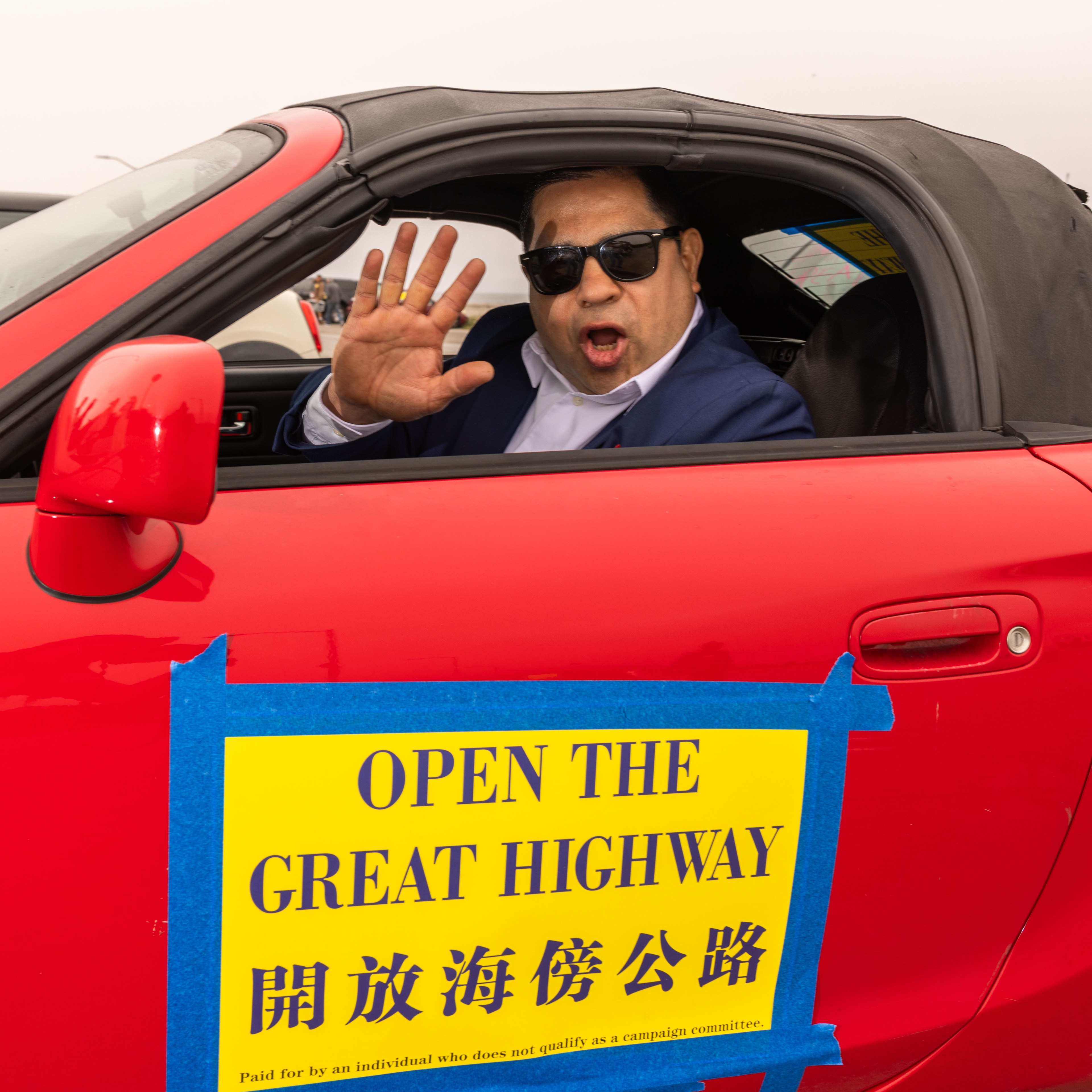 A man in sunglasses inside a red convertible, waving and shouting. The car has a sign reading "OPEN THE GREAT HIGHWAY" in English and Chinese.