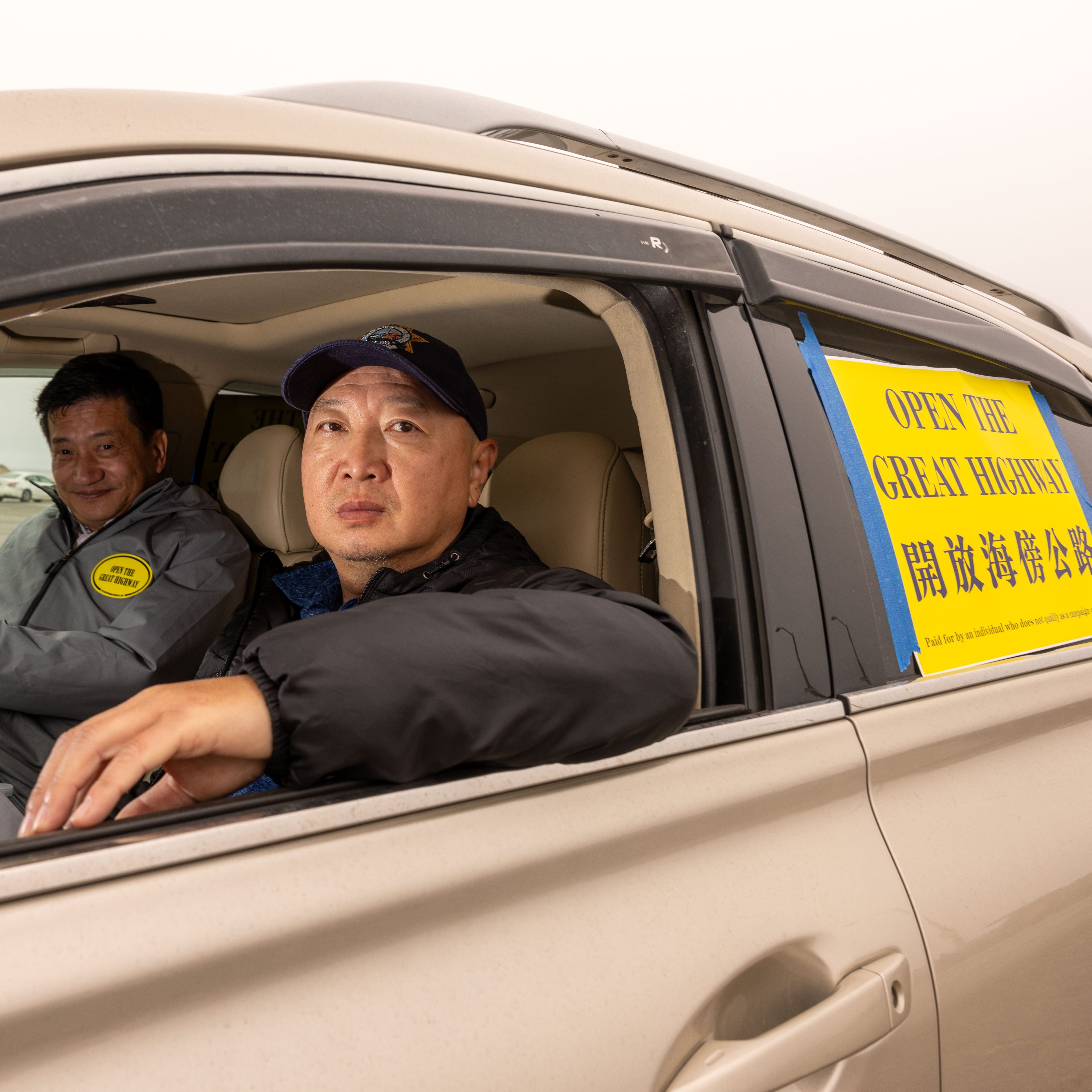 Two men are seen in a car with a sign on the door that reads &quot;OPEN THE GREAT HIGHWAY&quot; in both English and Chinese. The man in the passenger seat gazes seriously.