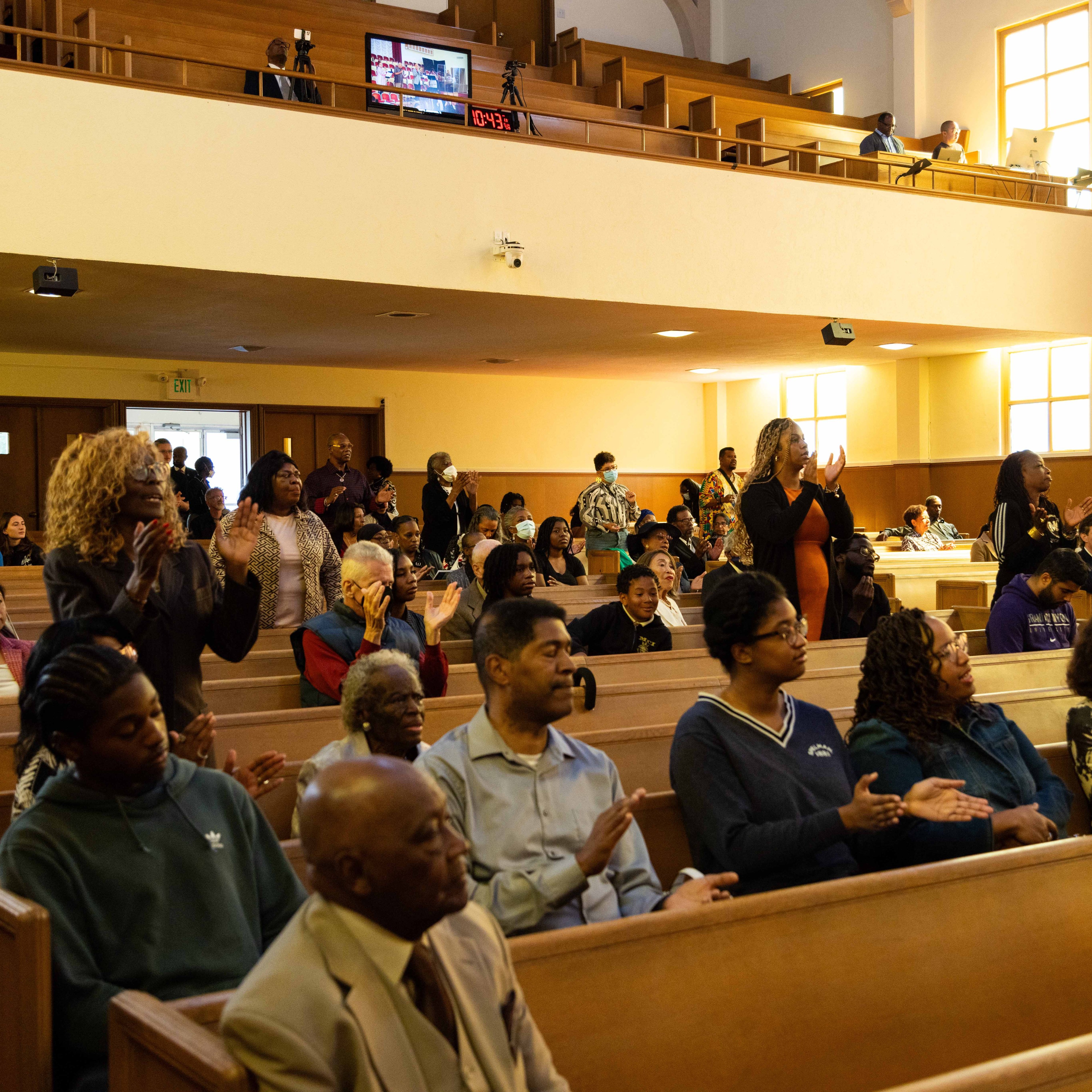 People sit and stand in a church, clapping and engaging in worship. The congregation is diverse, with individuals of various ages, and there's a balcony above.