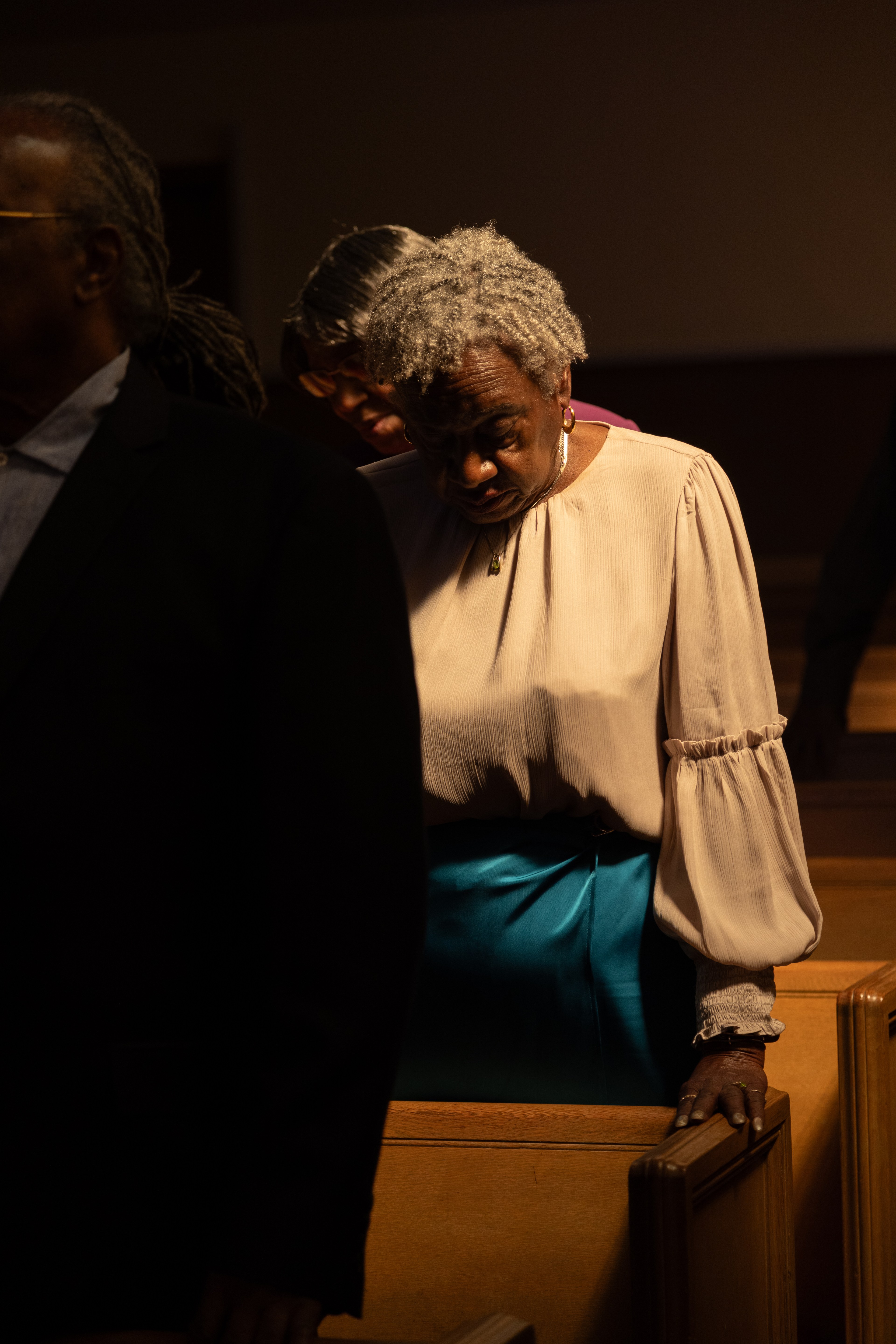 An elderly woman with gray hair is standing and leaning over a church pew, dressed in a light blouse and vibrant blue skirt. She appears to be in deep thought.