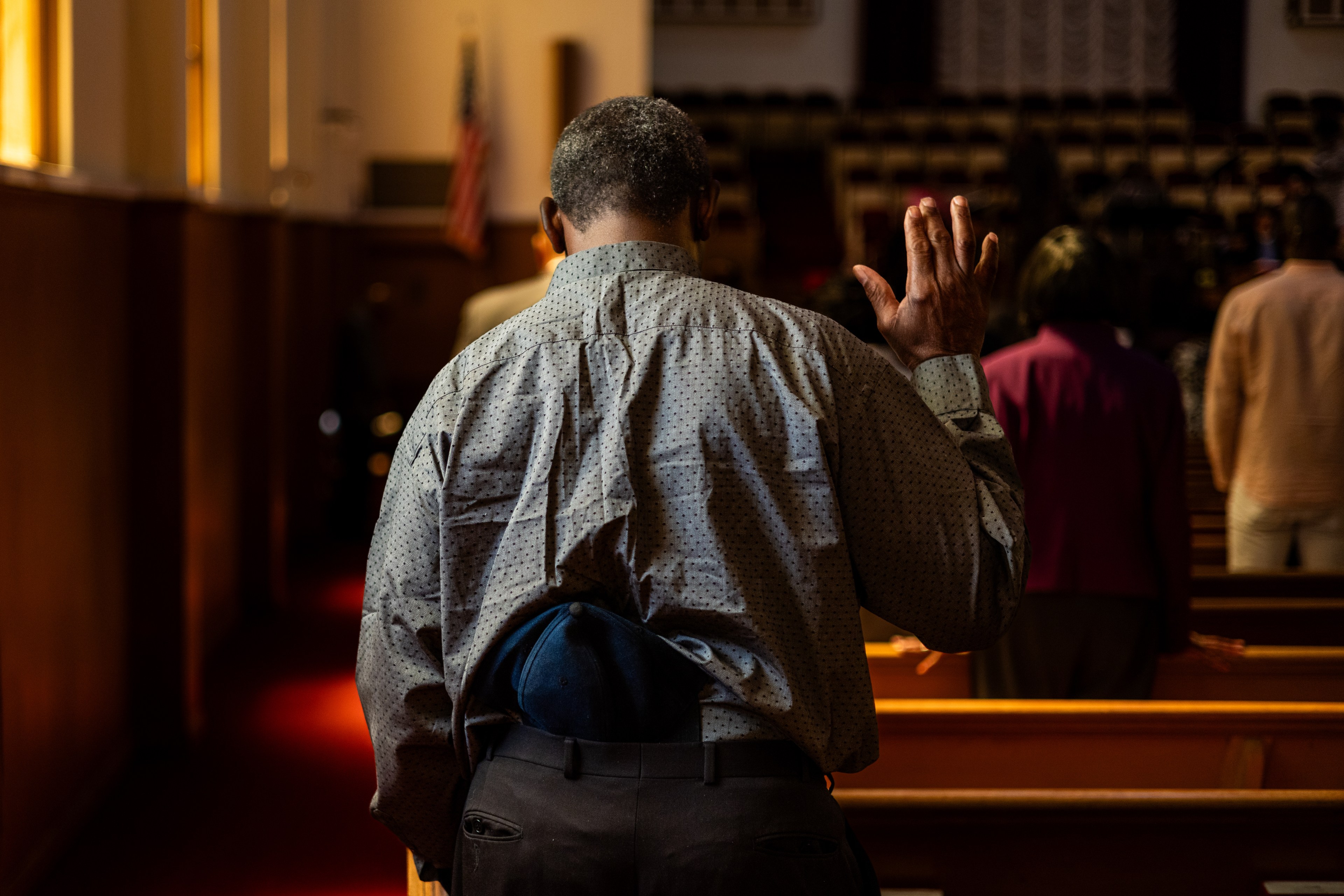 A man standing in a church pew raises his hand in worship, facing the front. He wears a dotted shirt and has a blue cap tucked into his waistband.