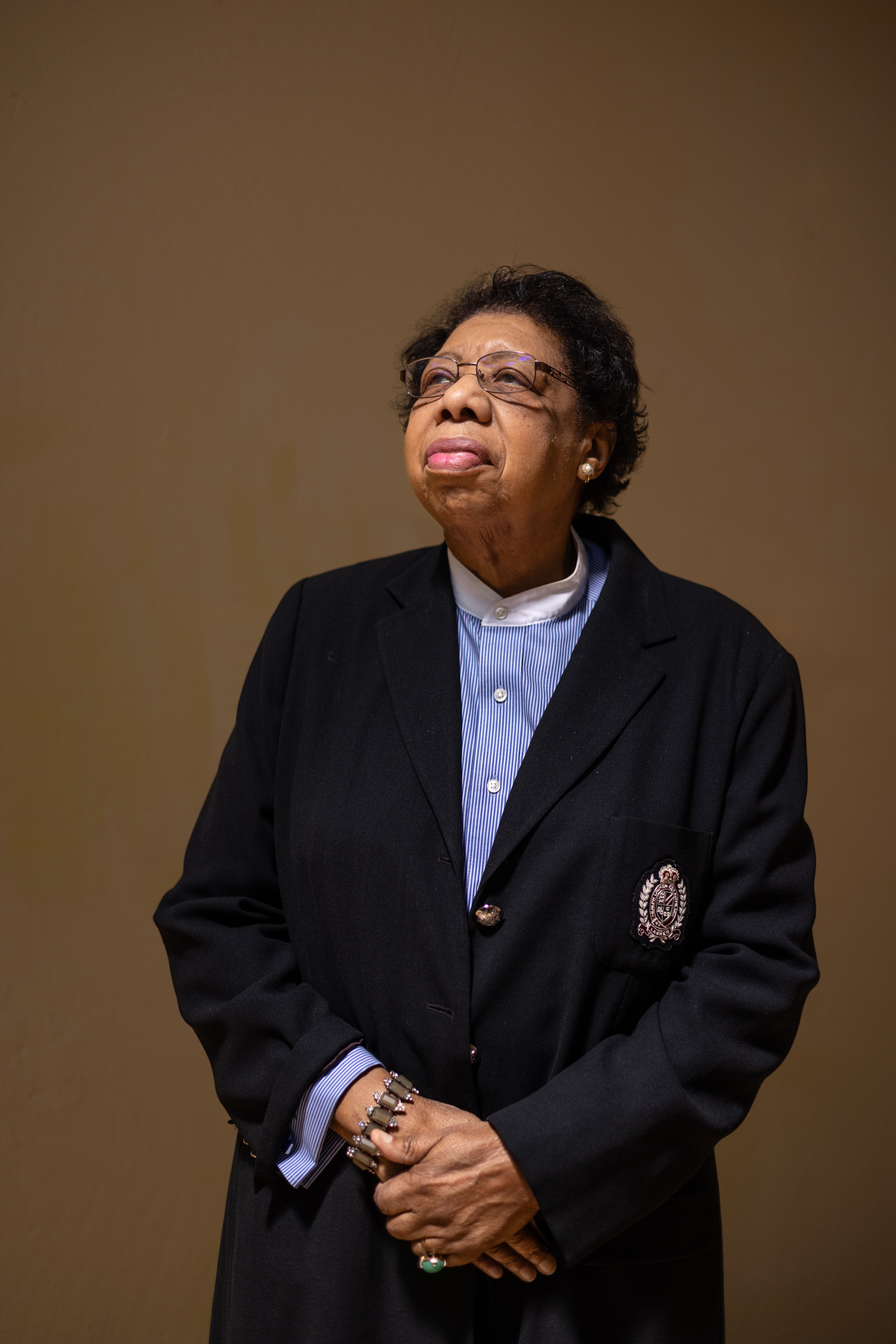 An elderly person wearing glasses and dressed in a dark blazer with a crest, a blue-striped shirt, and jewelry stands against a plain background, looking upward.