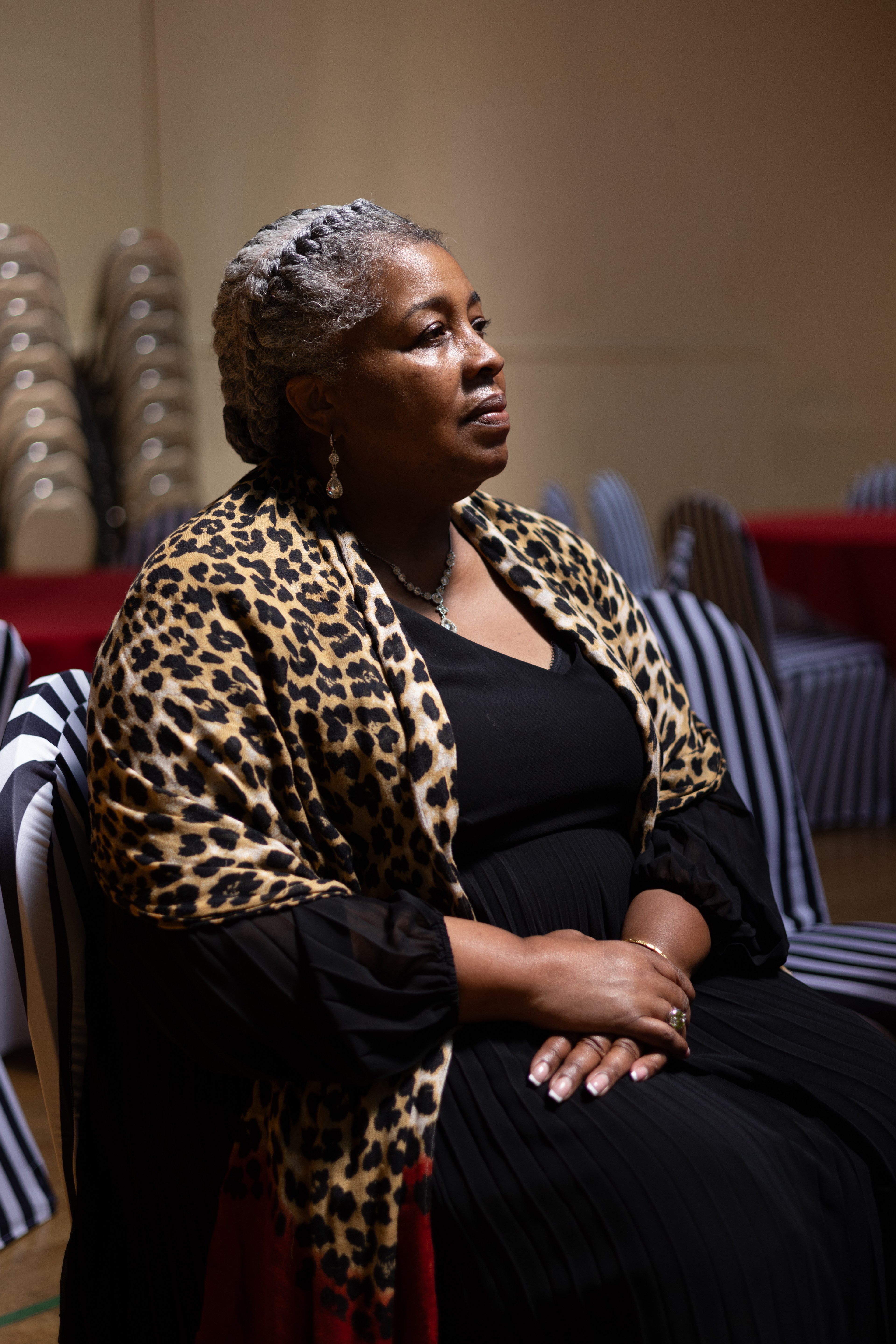 A woman with grey braids, wearing a black dress and leopard print shawl, sits with hands folded in a dimly lit room with striped chair covers and stacked chairs.