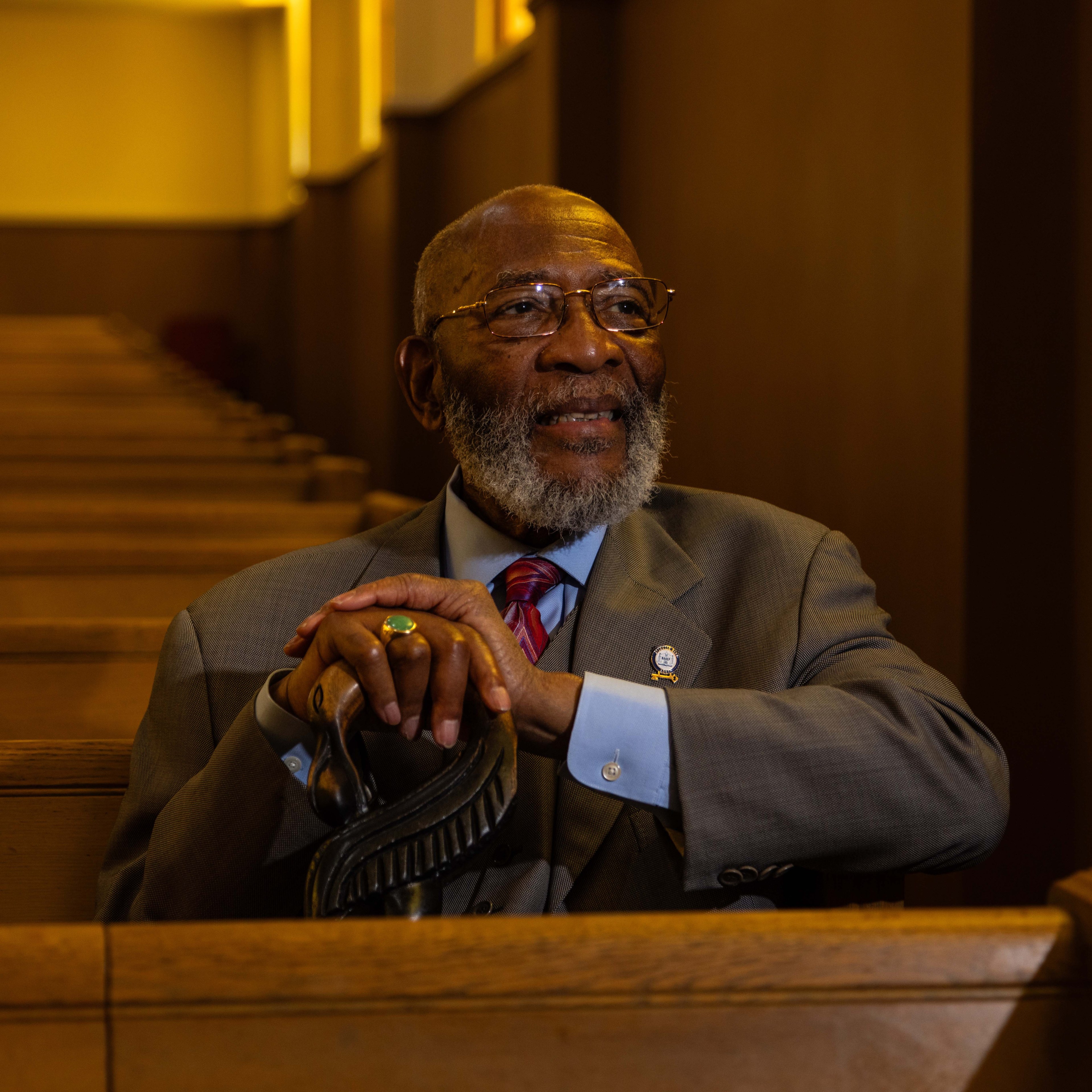 A man with a gray beard wearing a suit and glasses sits in a wooden pew, smiling, resting his hands on a cane, with warm lighting illuminating the background.