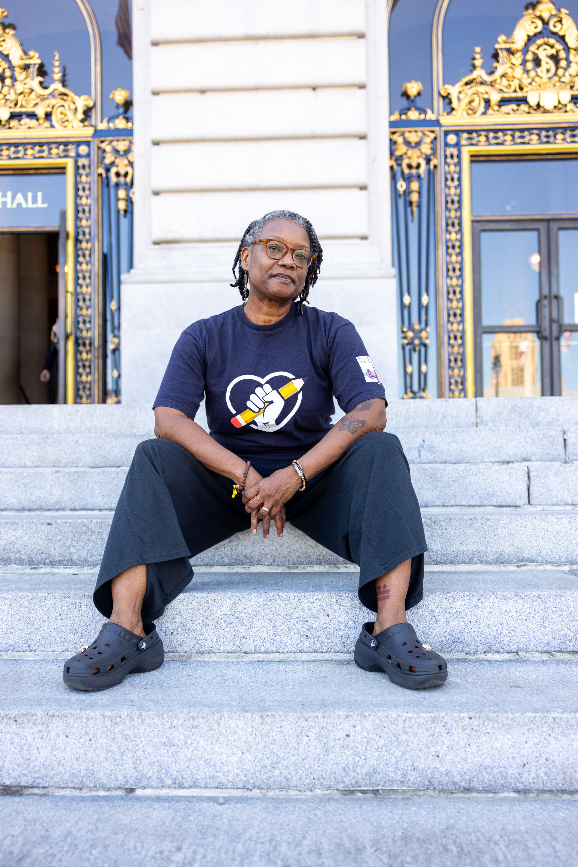 A person with dreadlocks, wearing glasses, a navy shirt with a fist and pencil logo, dark pants, and black Crocs, sits on ornate building steps with golden details.