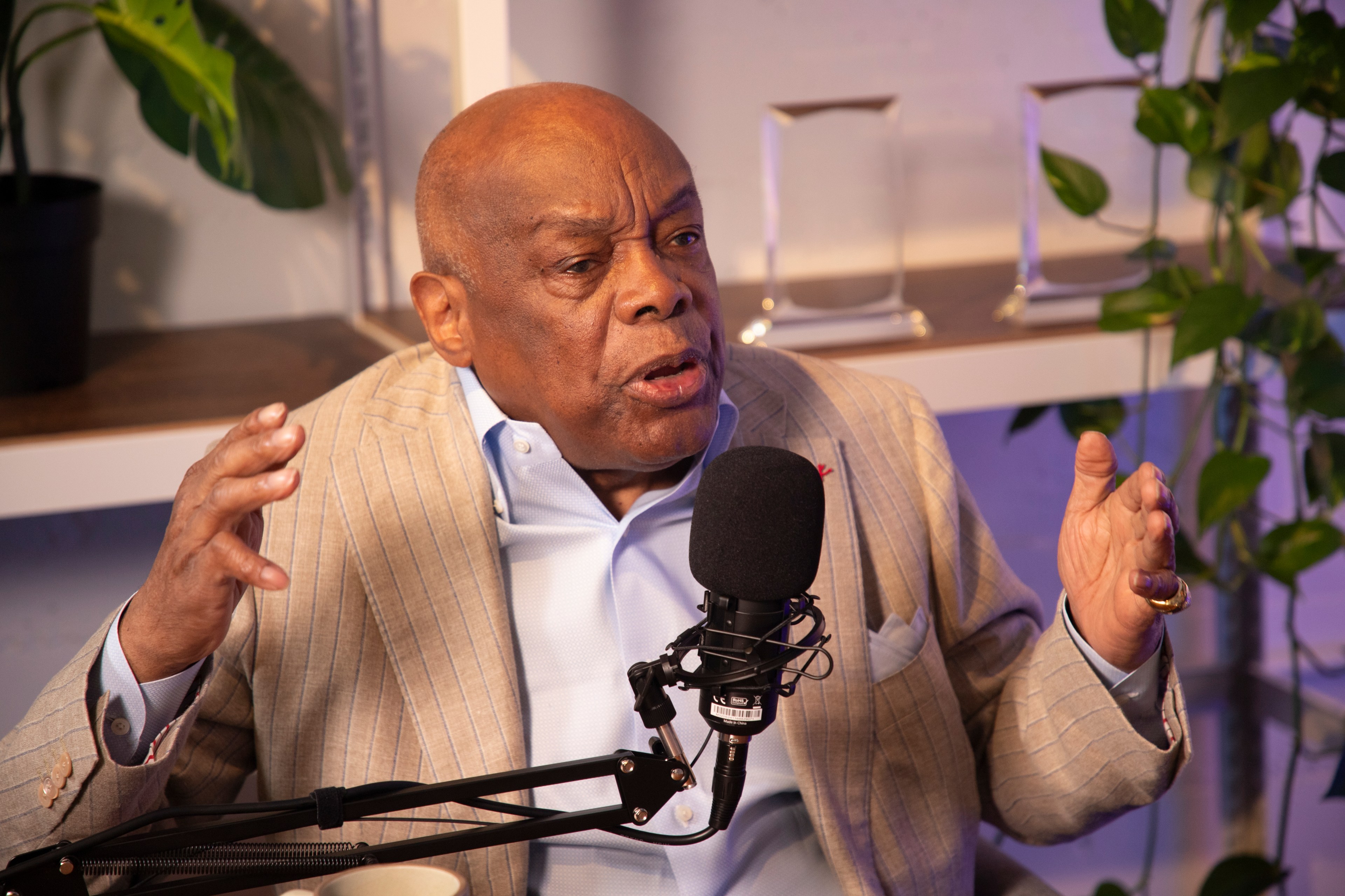 An elderly man in a beige pinstripe suit speaks passionately into a microphone, gesturing with his hands. He sits before bookshelves adorned with plants.