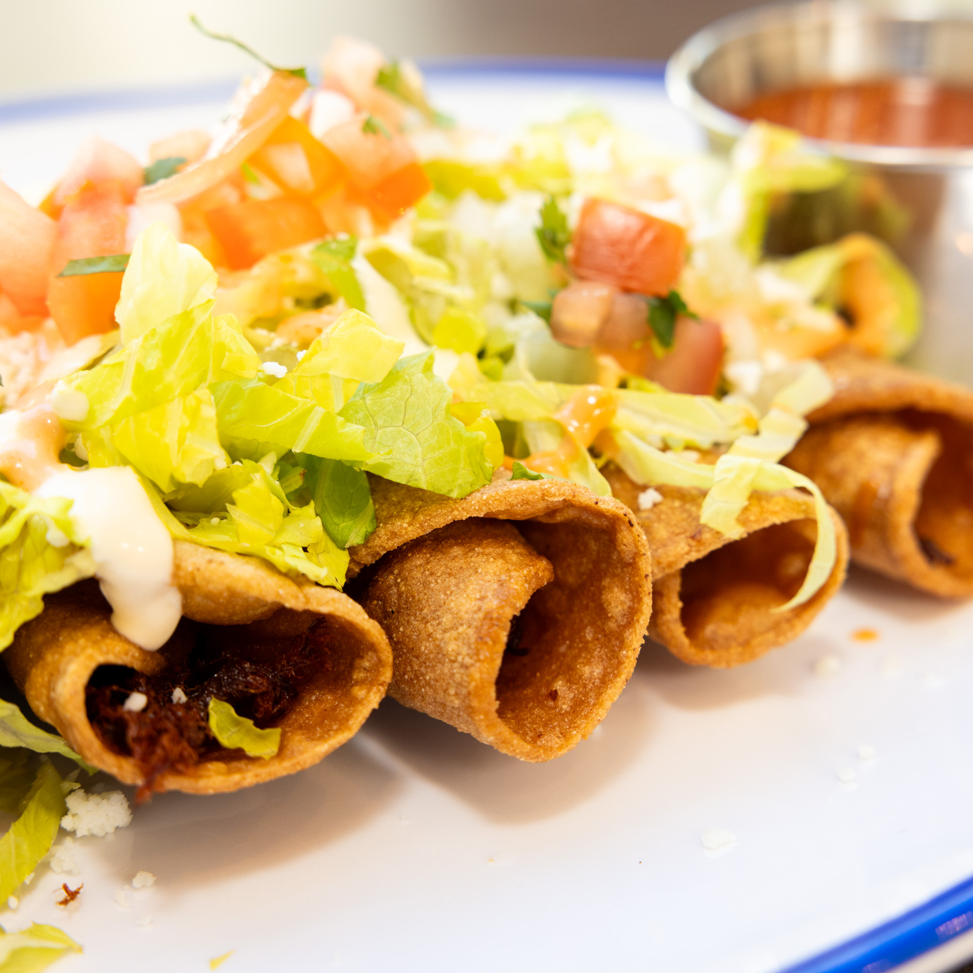 Three crispy taquitos topped with chopped lettuce, tomatoes, cheese, and drizzled sauce on a white plate with a side of red salsa in a small metal cup.