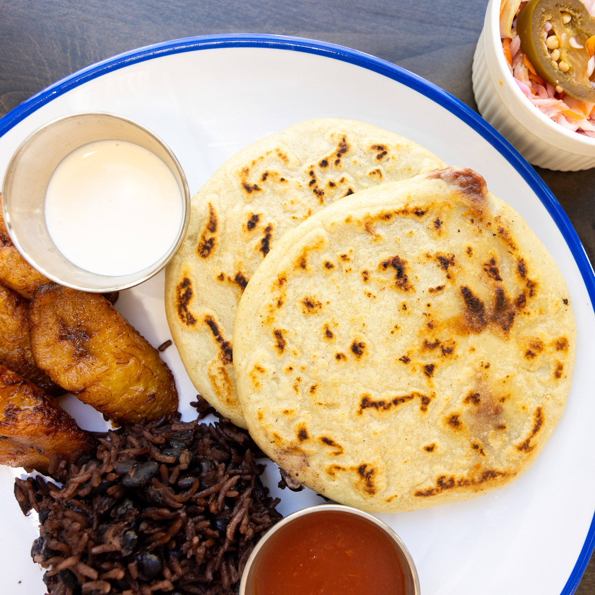 The image shows a plate with two browned corn pupusas, black beans and rice, fried plantains, and sides of white sauce, red sauce, and pickled vegetables.