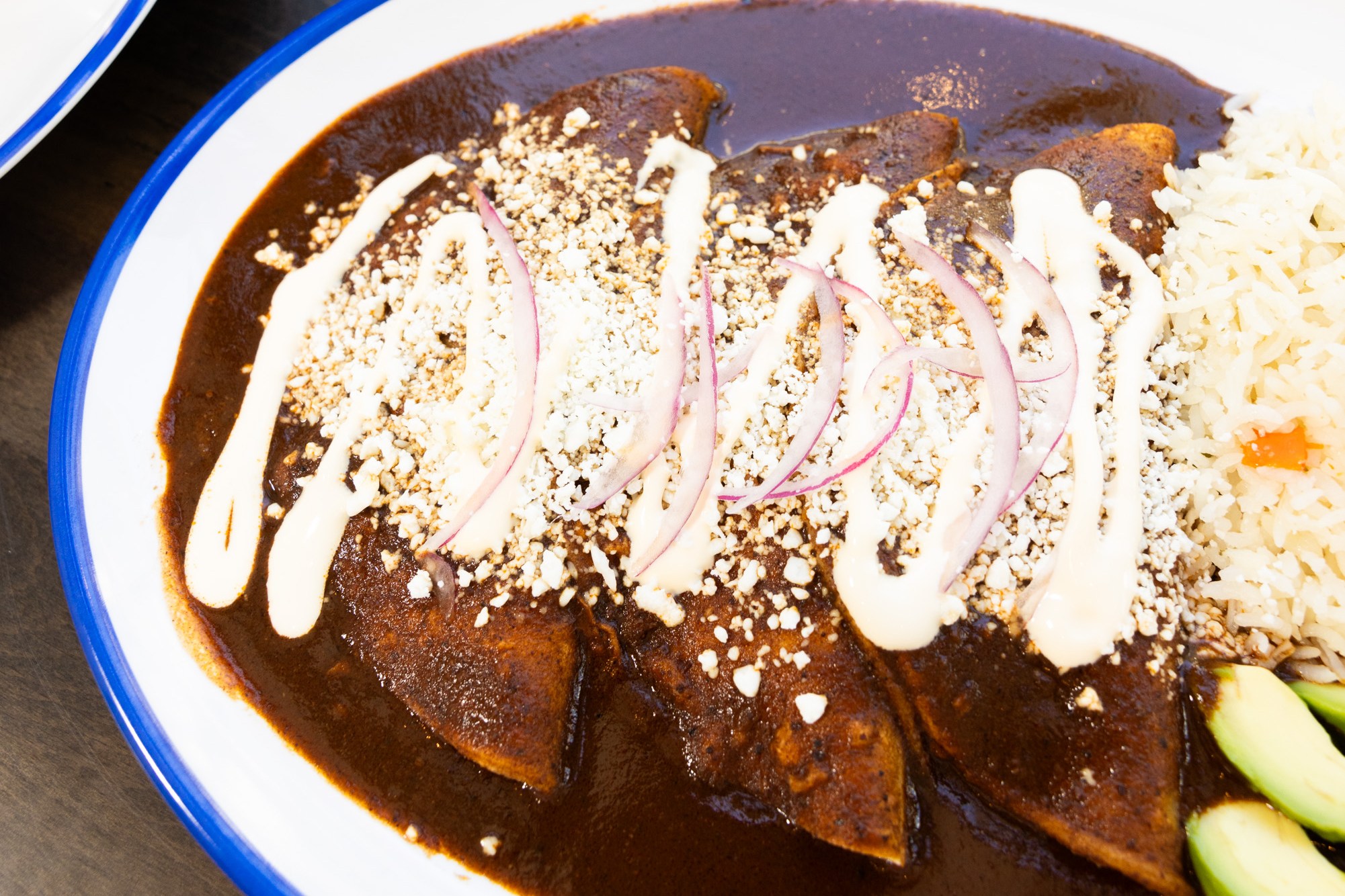 The image shows two oval plates of Mexican food, one with enchiladas covered in mole sauce, crema, and sliced onions, the other with crispy flautas topped with lettuce, tomato, and crema.