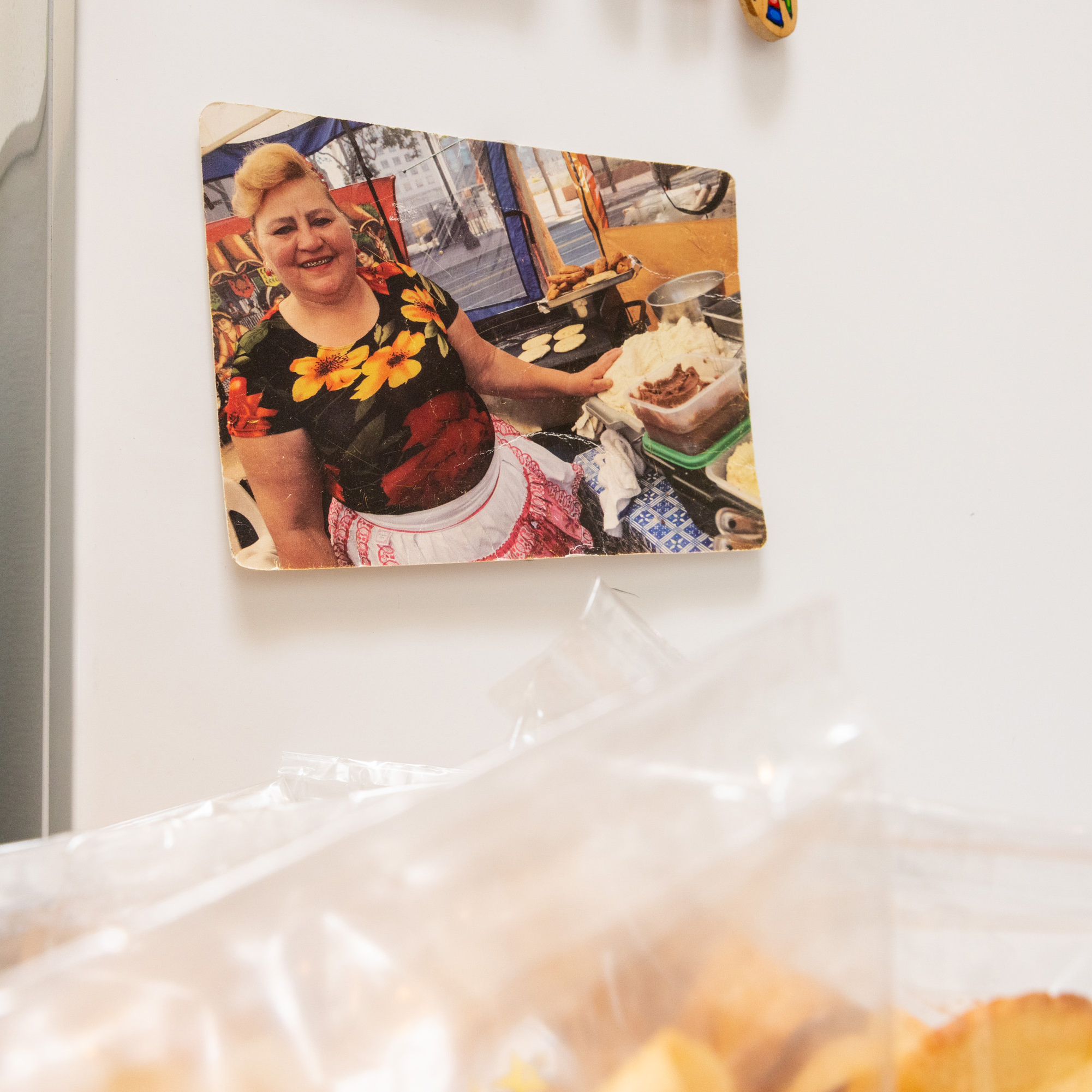 A smiling woman with blonde hair, wearing a floral top and apron, stands at a street food stall with cooking equipment and ingredients visible.