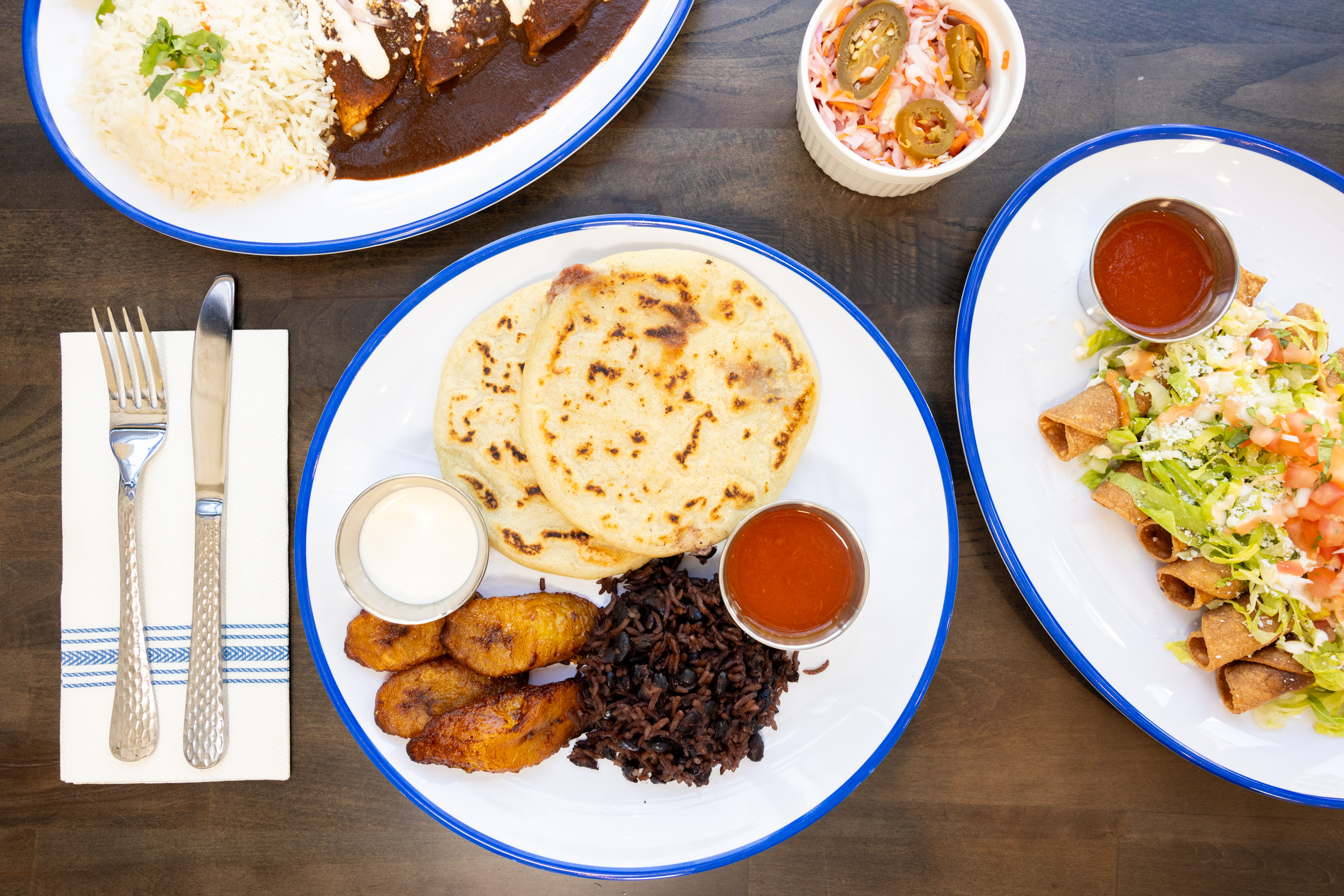 The image shows a table set with various dishes, including pupusas, fried plantains, rice, black beans, enchiladas, and taquitos, accompanied by dipping sauces.