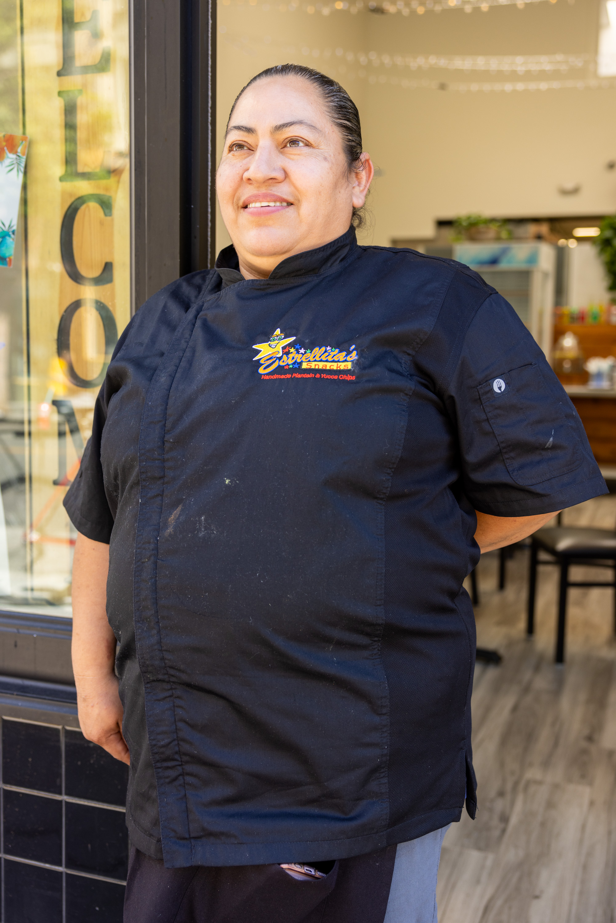 A chef stands smiling beside a &quot;Welcome&quot; sign in a doorway. She wears a black uniform with an embroidered logo and a clean, well-lit space is visible behind her.