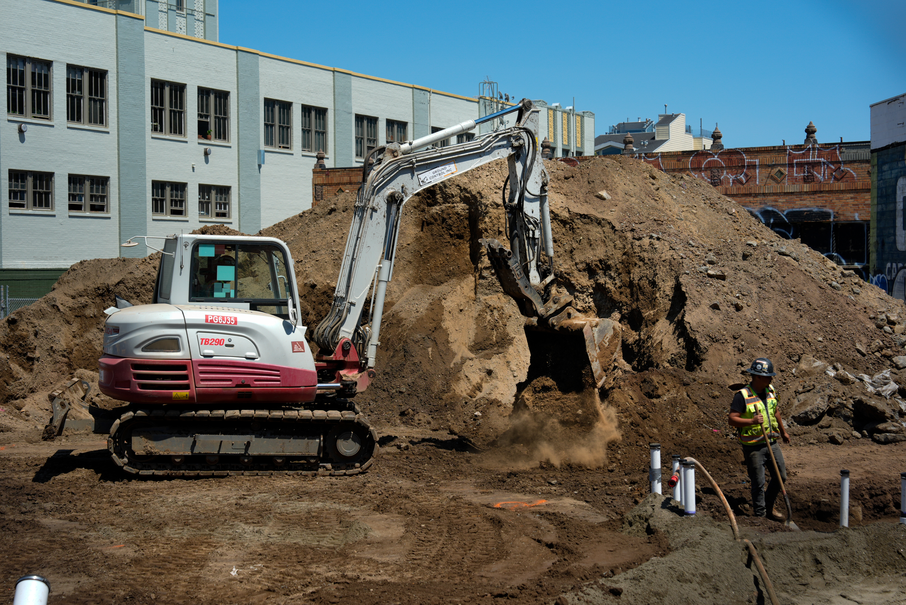 A construction site with a white and red excavator moving dirt near a large pile. A worker in a yellow vest and hard hat stands nearby, holding a shovel.