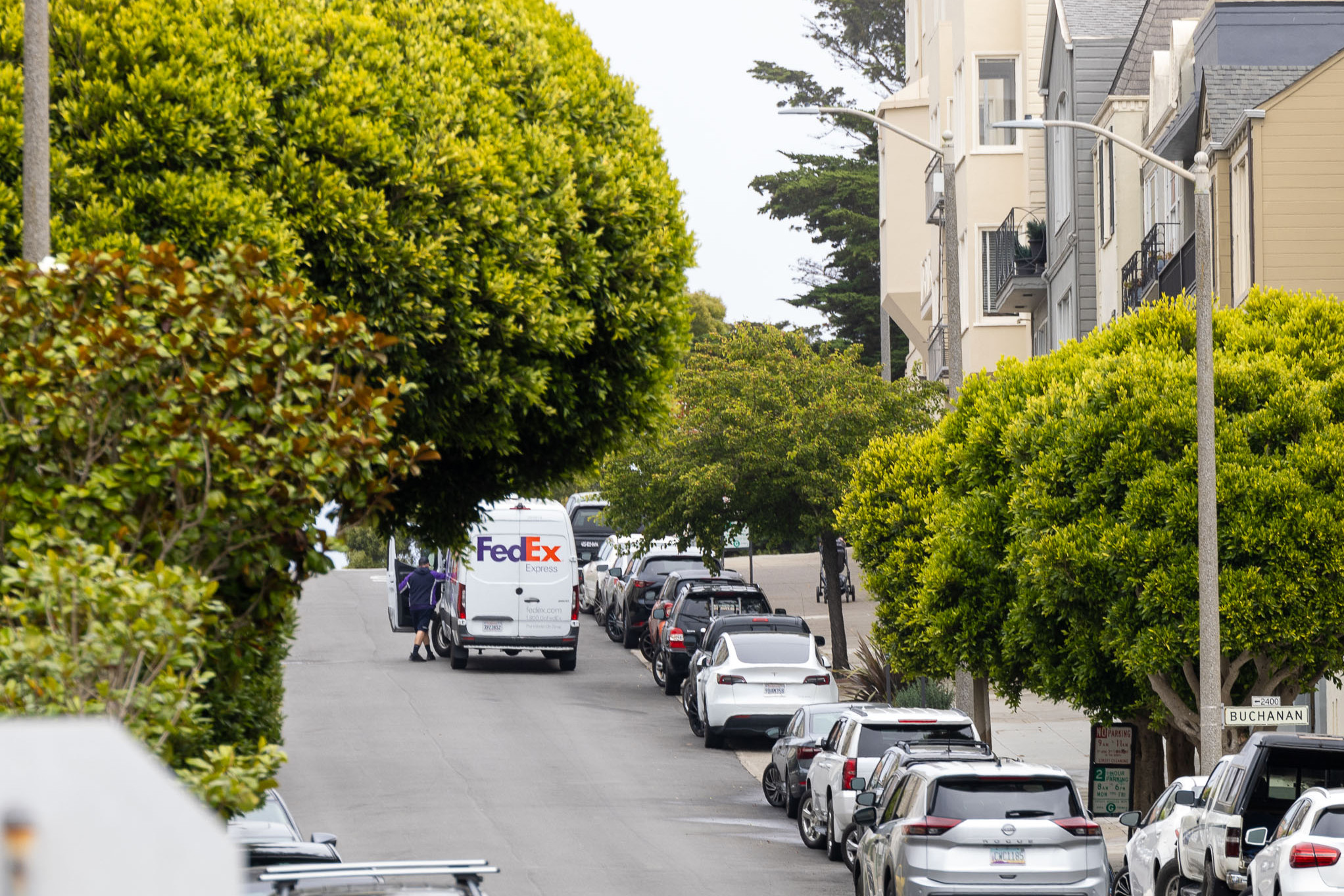 The image shows a residential street lined with trees and parked cars. A FedEx truck is stopped on the side, with a delivery person visible at the truck's side door.