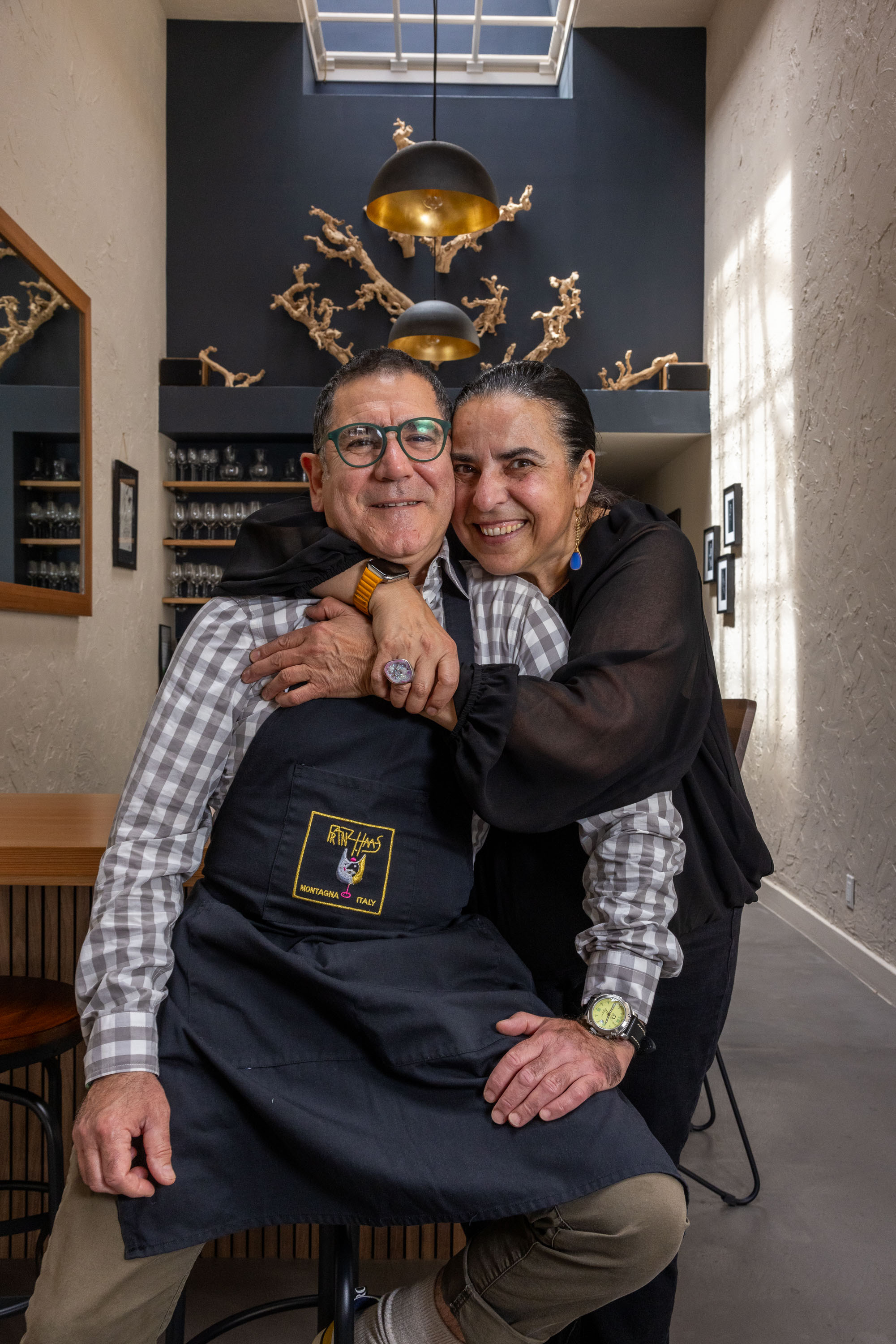 A smiling couple embraces in a stylish, modern room featuring a skylight, black wall, golden sculptural decor, and shelves of glassware. The man wears glasses and an apron.