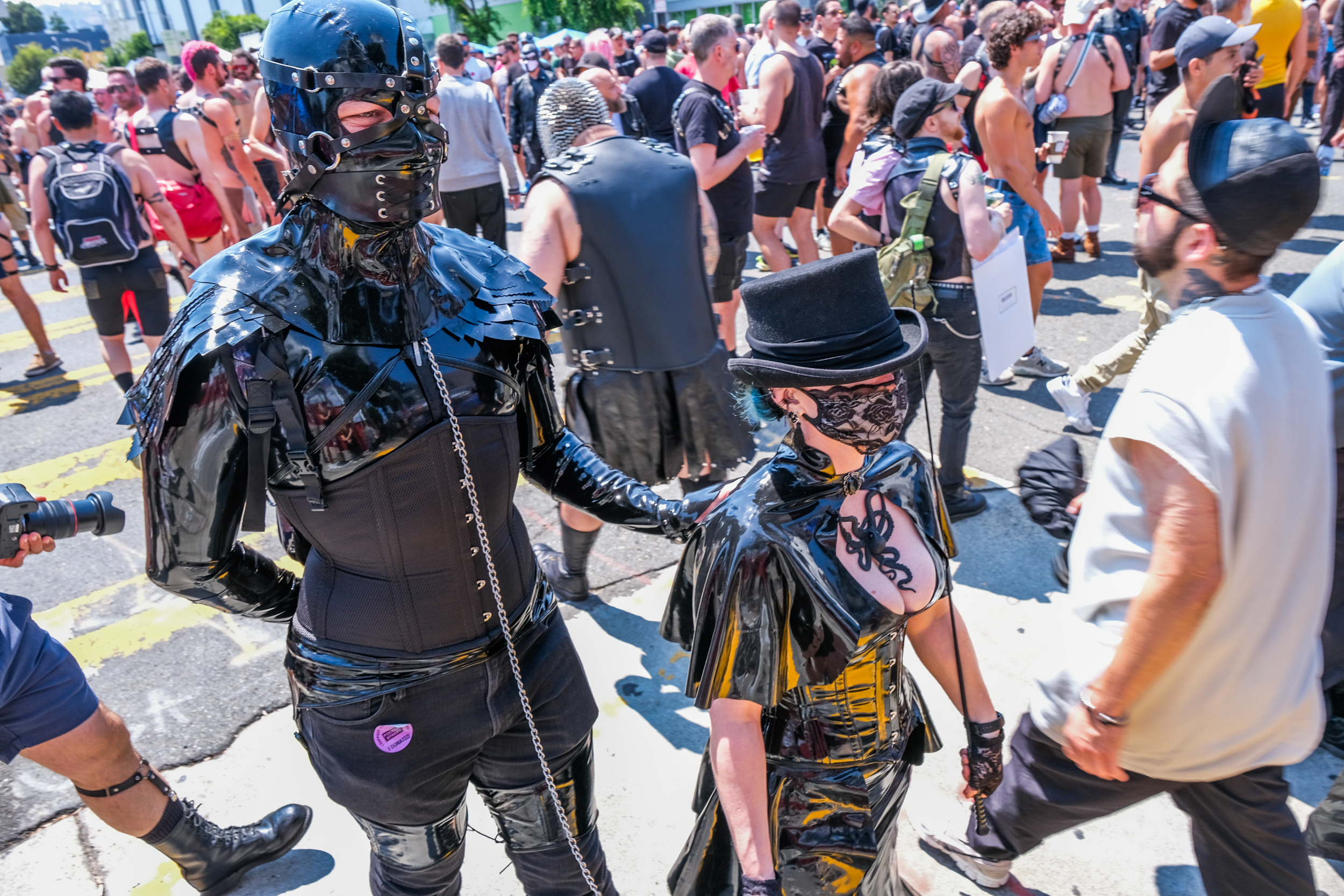 Two individuals in black, glossy, fetish attire with masks and chains walk among a crowd, one appears to be leading the other. Many onlookers are dressed similarly.