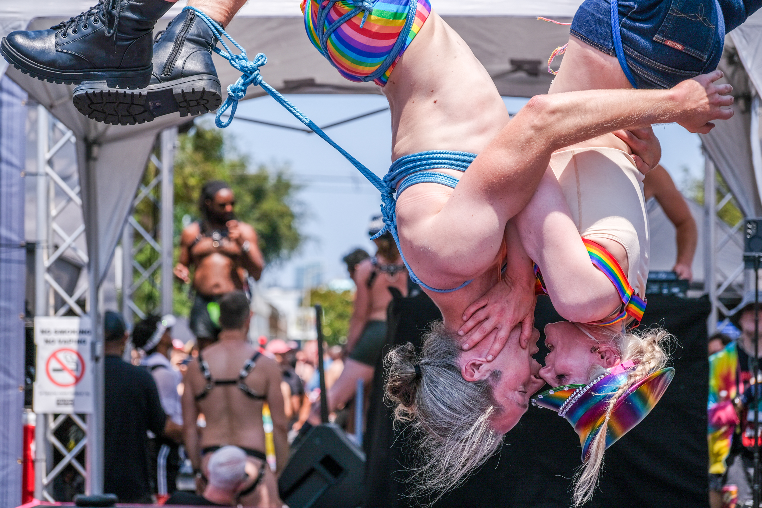 Two people are suspended upside down, embracing each other. They are wearing colorful rainbow attire and surrounded by a crowd, possibly at a festival or event.