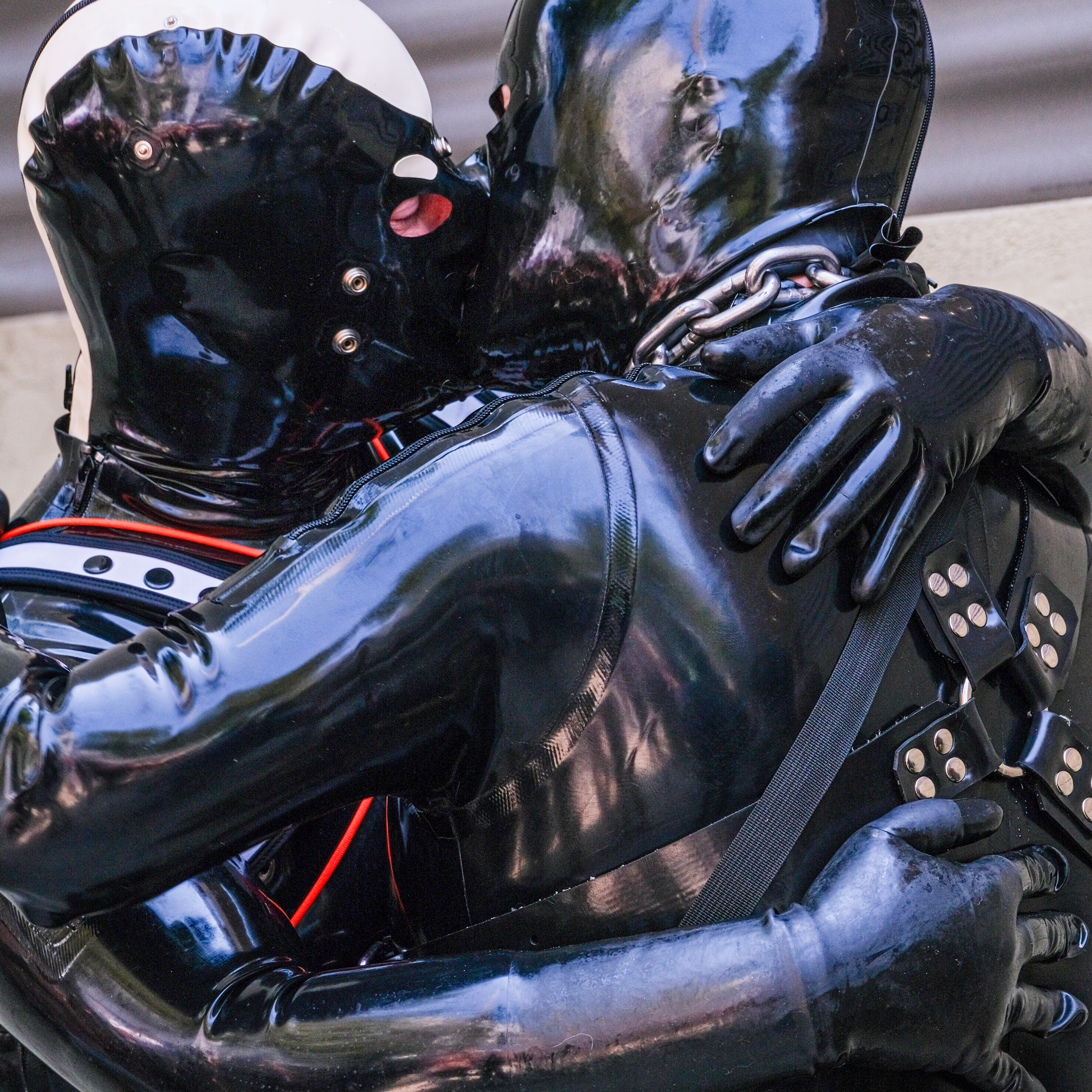 Two individuals, clad in shiny black latex suits and masks, embrace closely, with one wearing a white accent on their mask and the other sporting a harness with metal details.