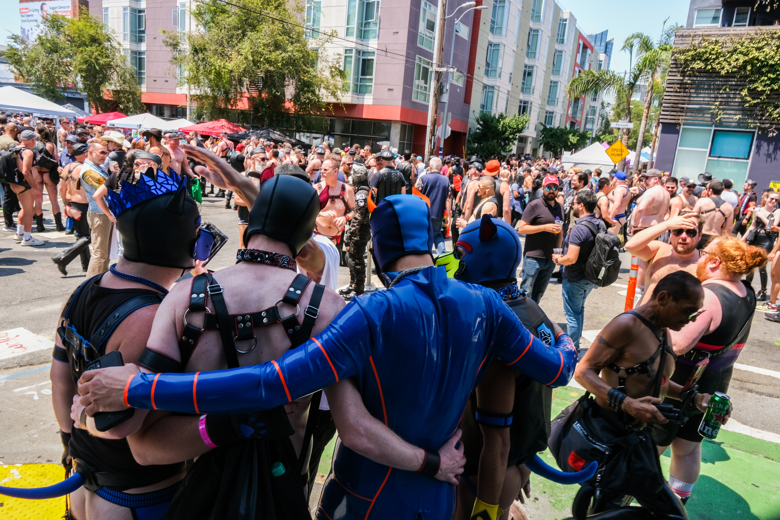 A group of people in colorful, leather outfits and masks embrace in the foreground, with a large crowd similarly dressed at an outdoor event in an urban setting.