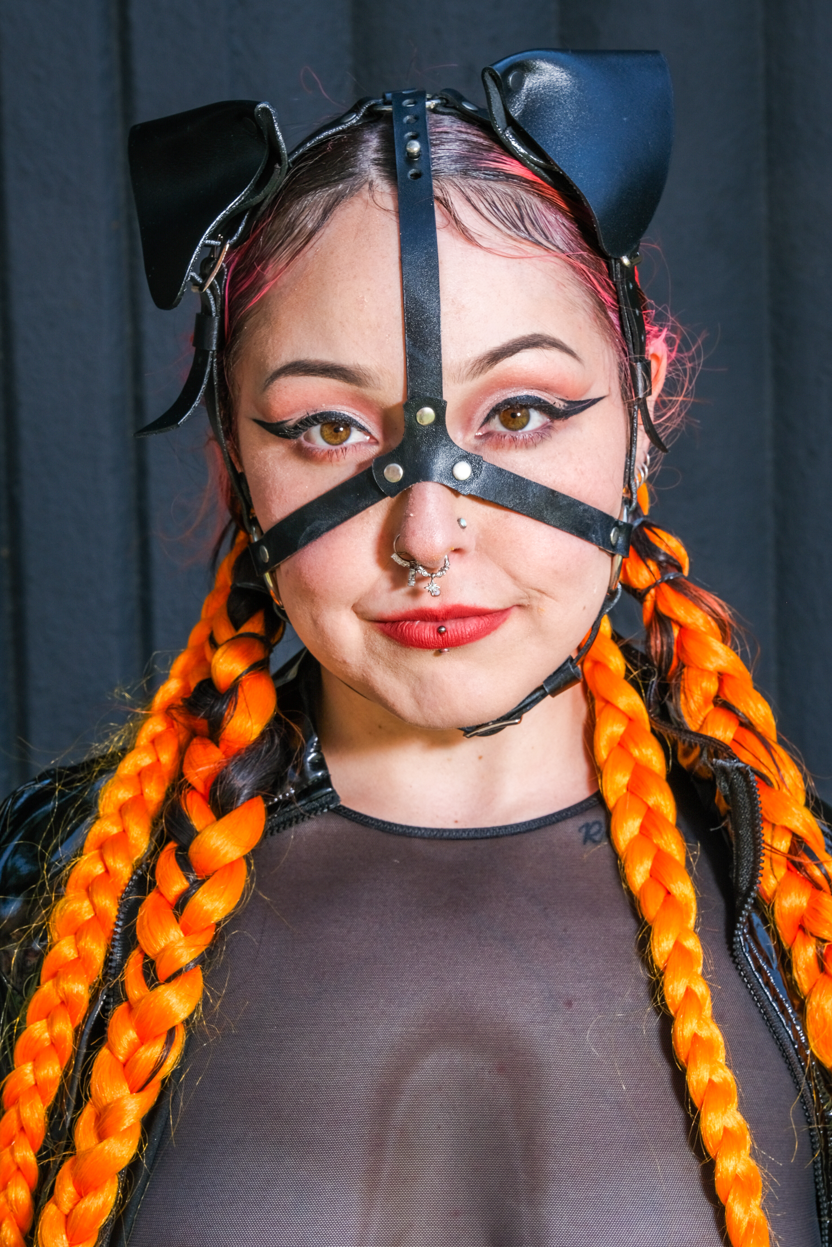 A person with bright orange braided hair is wearing a sheer black outfit and a black leather head harness with cat ears, showcasing facial piercings and red lipstick.