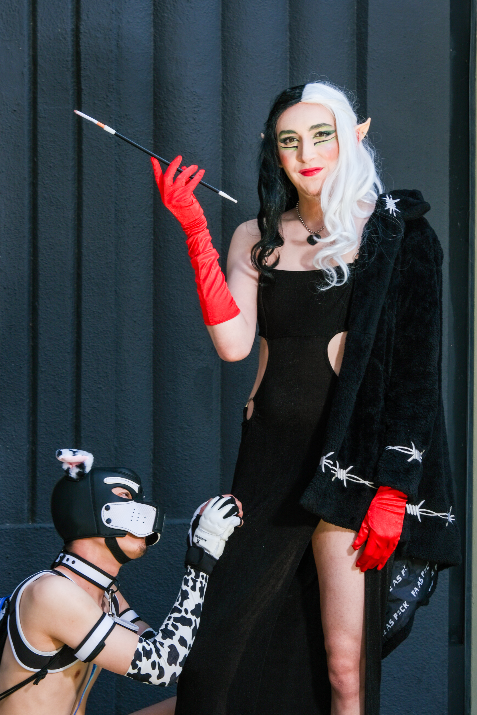 A person dressed in black and red gloves, with a black-and-white wig, stands holding a long cigarette holder. Another person, wearing a dog mask, kneels and looks up at them.