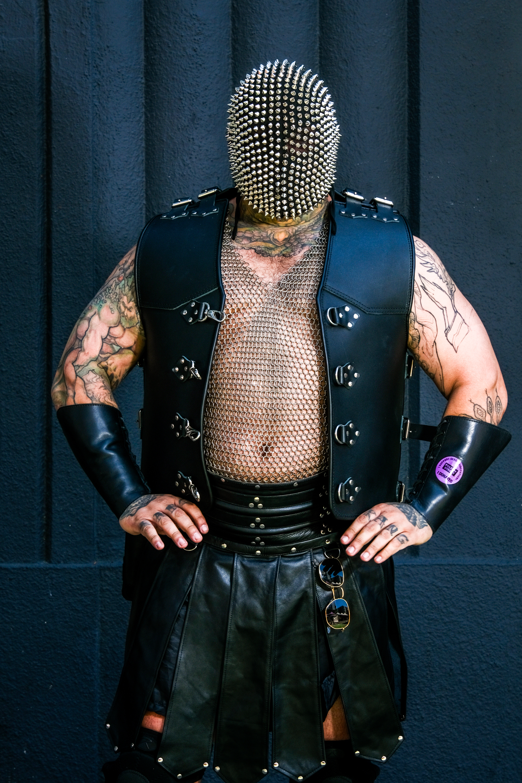 A person in spiked headgear, leather vest, mesh top, leather skirt, and arm guards with tattoos and sunglasses.