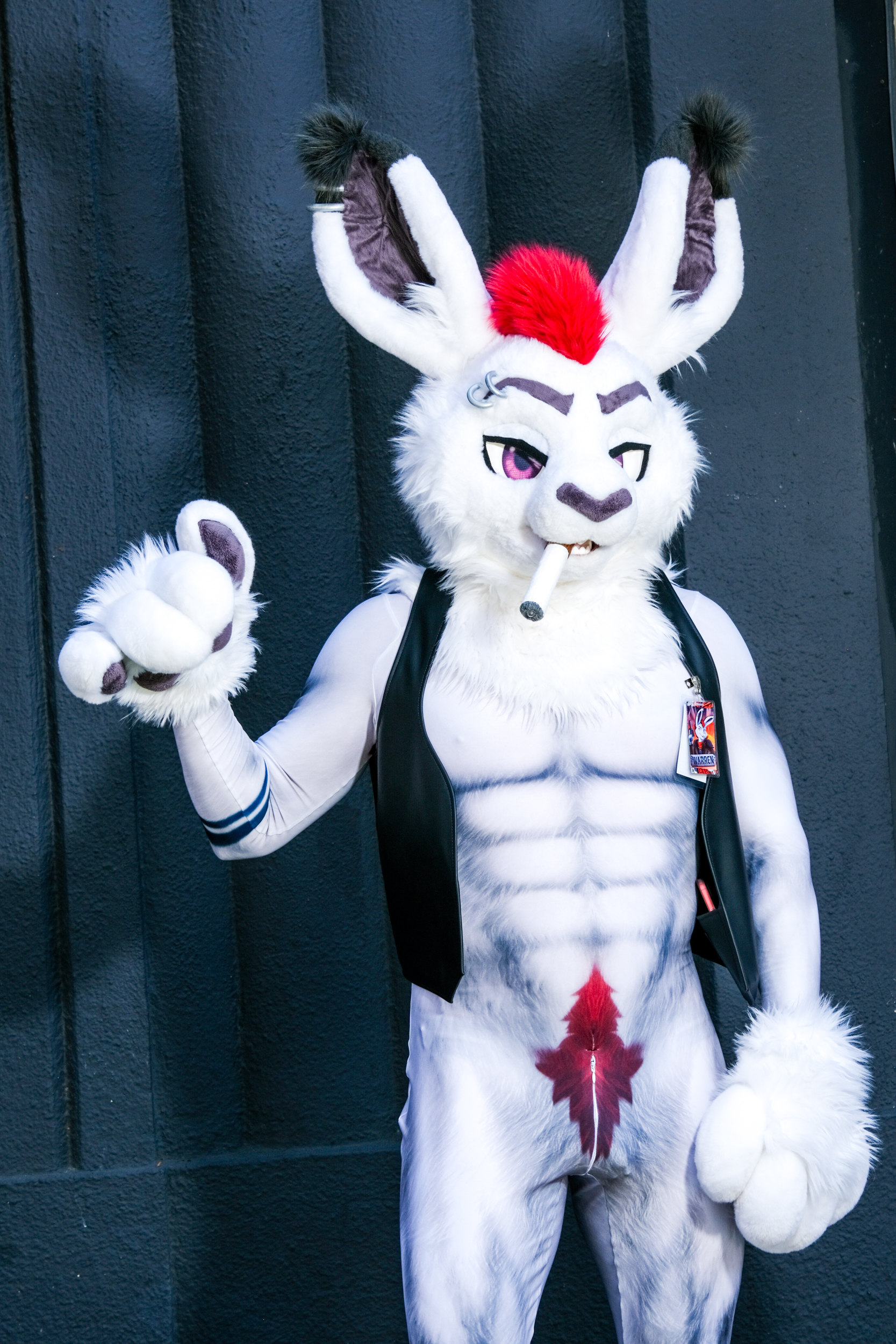 A costumed character resembling a muscular white rabbit with a red mohawk, wearing a black vest, showing off a pose with a cigarette in its mouth against a dark background.