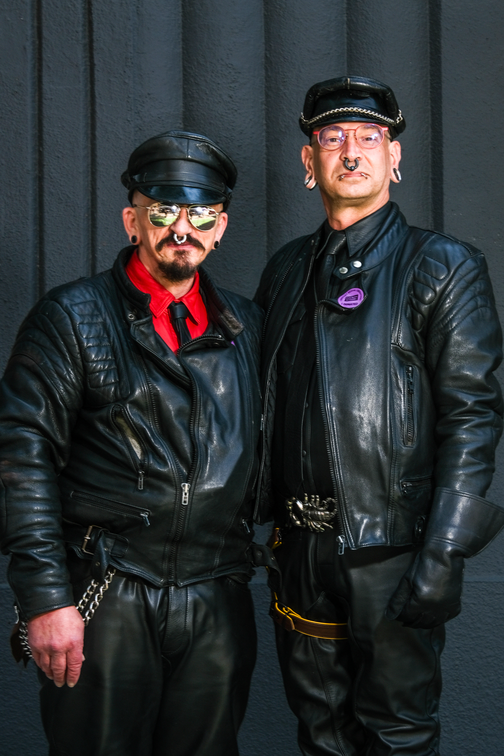 Two individuals dressed in black leather jackets, pants, and hats stand side by side. Both have nose piercings and glasses, and one wears a red shirt and tie.