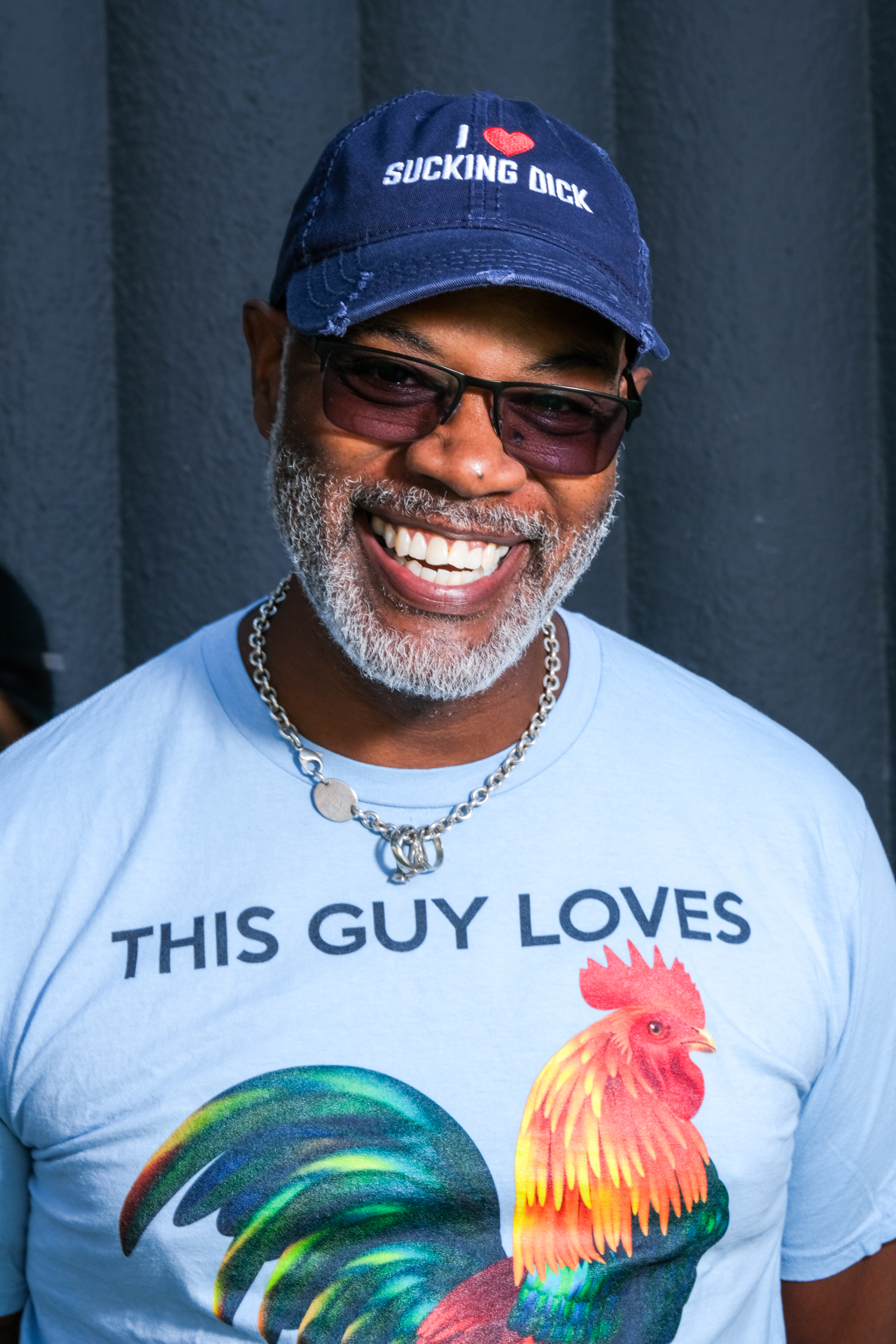 A smiling man wears a cap with an explicit message and a shirt with a colorful rooster and the text &quot;THIS GUY LOVES.&quot; He also has a chain necklace and glasses.