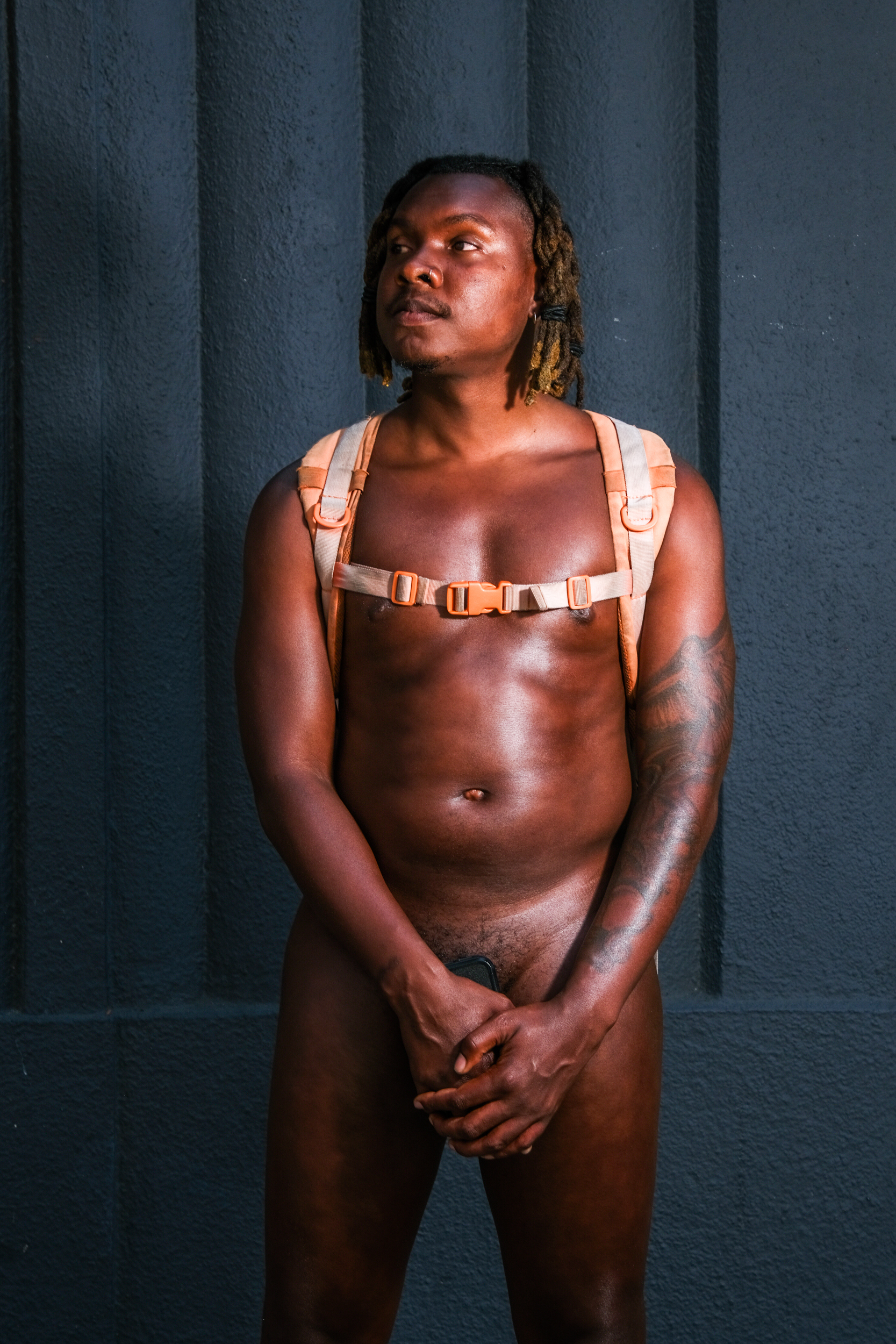 A nude person with an orange backpack stands against a dark textured wall, hands clasped strategically in front. Their hair is styled in short dreadlocks.